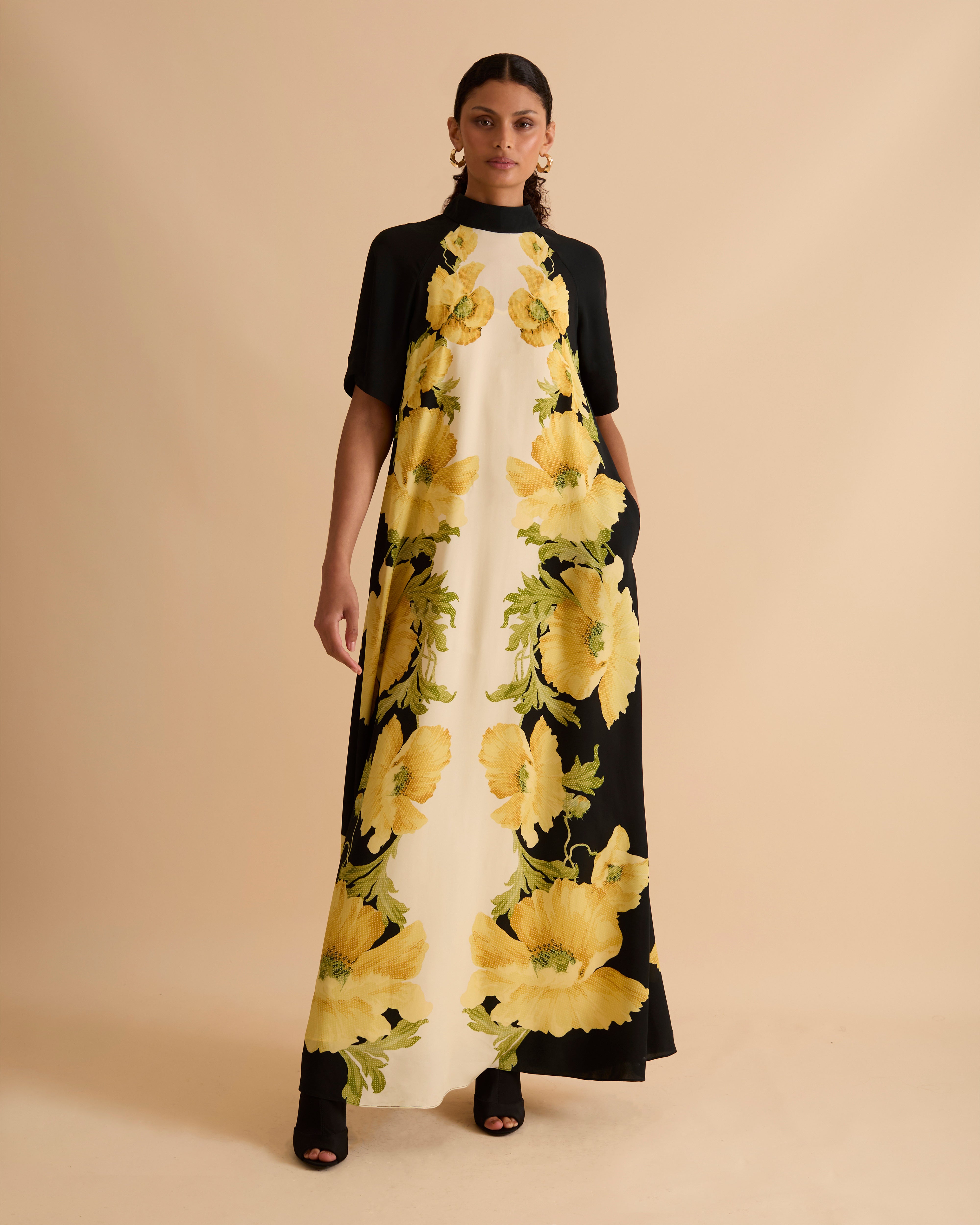 Model wearing Florere black and ivory maxi dress with bold yellow floral print, high neckline, short sleeves, and side pockets, styled with gold hoop earrings and black open-toe boots, against a neutral beige background