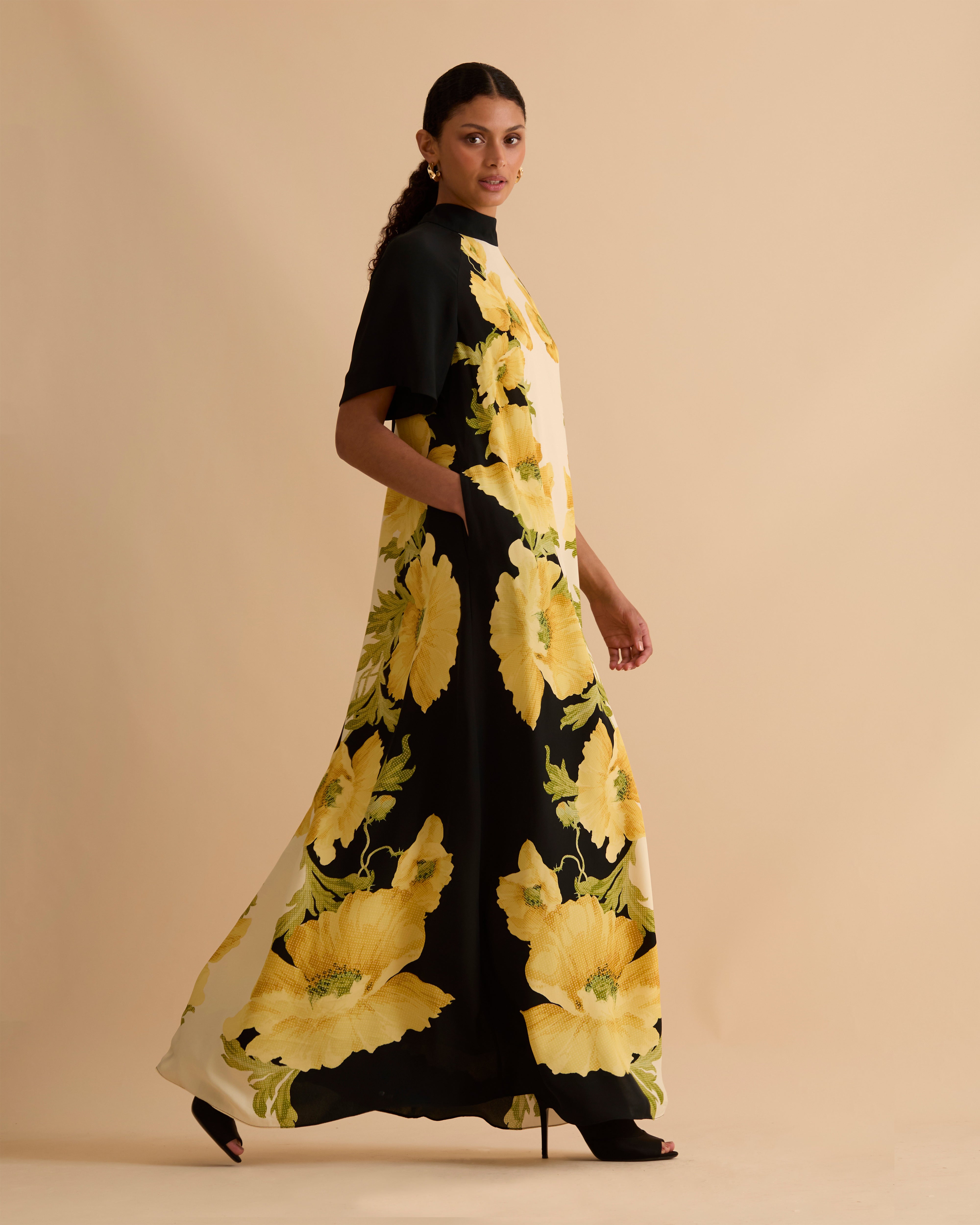 Model wearing Florere black and ivory maxi dress with bold yellow floral print, high neckline, short sleeves, and side pockets, styled with gold hoop earrings and black open-toe boots, against a neutral beige background