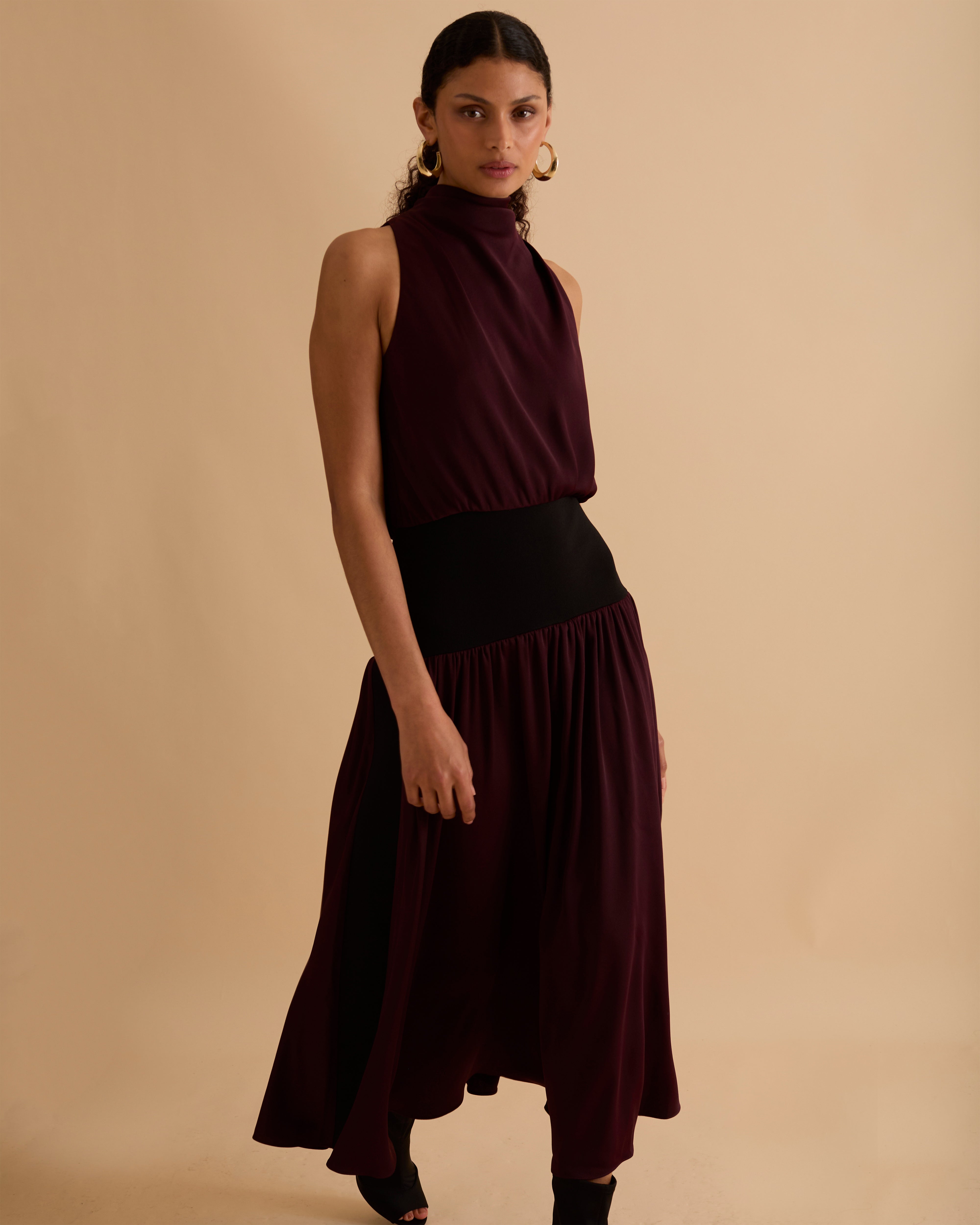 Model wearing Florere sleeveless burgundy midi dress with high neckline, black waistband detail, and flowy A-line silhouette, styled with gold hoop earrings and black open-toe heels, standing against a beige background