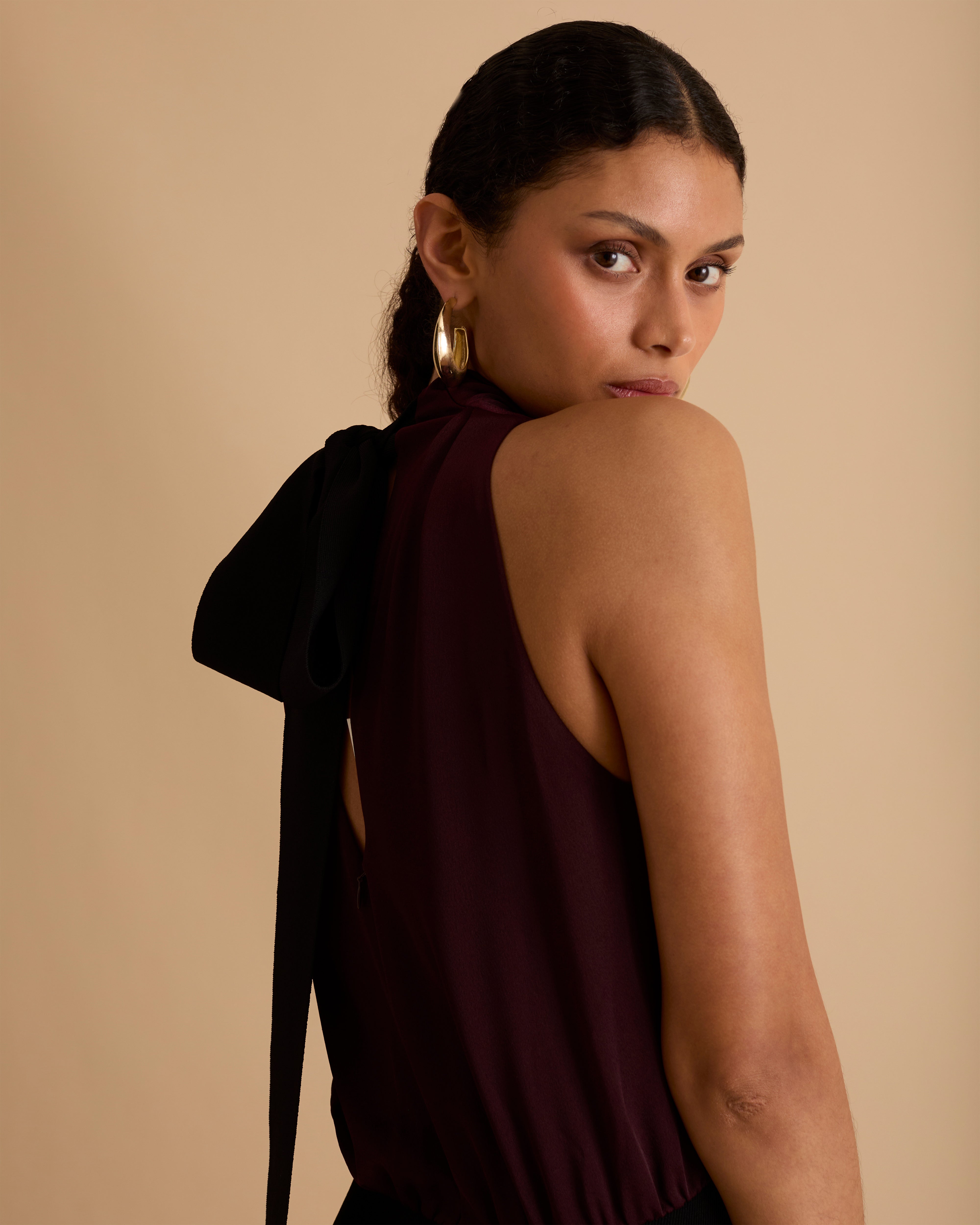 Model wearing Florere sleeveless burgundy midi dress with high neckline, black waistband detail, and flowy A-line silhouette, styled with gold hoop earrings and black open-toe heels, standing against a beige background