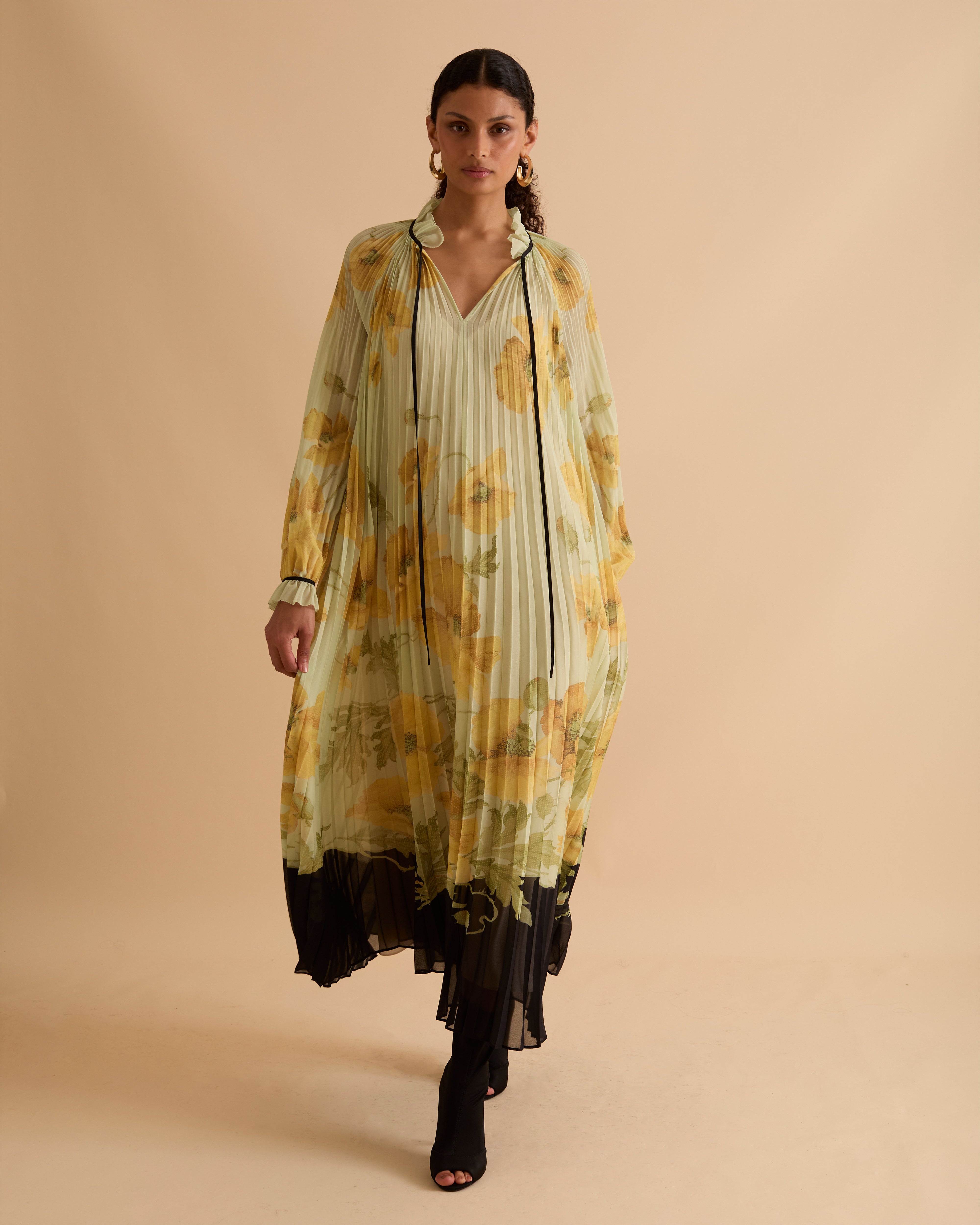 Model wearing Florere pleated floral maxi dress with yellow poppy print, sheer long sleeves, high neckline with ties and black hem, styled with gold hoop earrings and black open-toe boots against a beige background