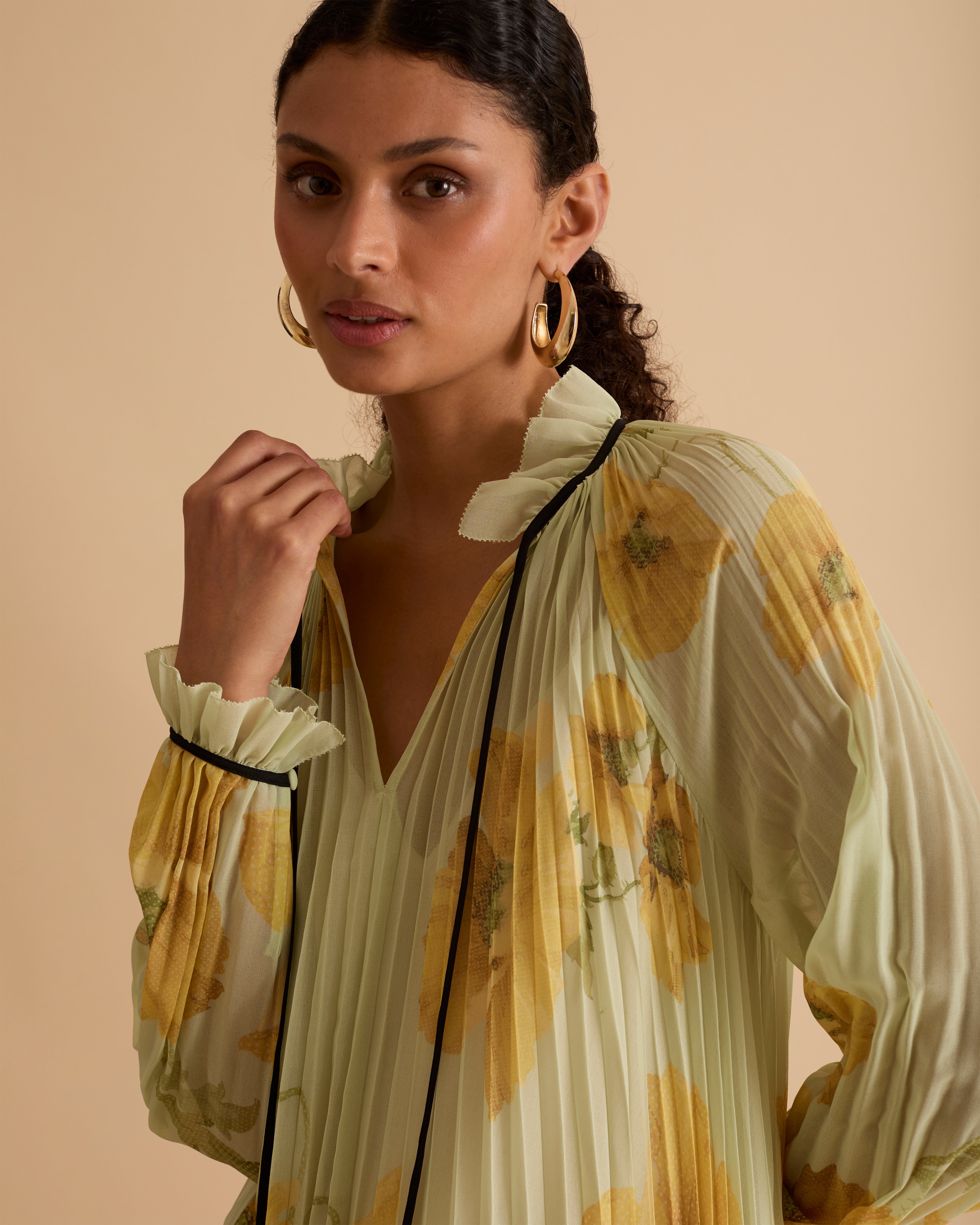 Model wearing Florere pleated floral maxi dress with yellow poppy print, sheer long sleeves, high neckline with ties and black hem, styled with gold hoop earrings and black open-toe boots against a beige background