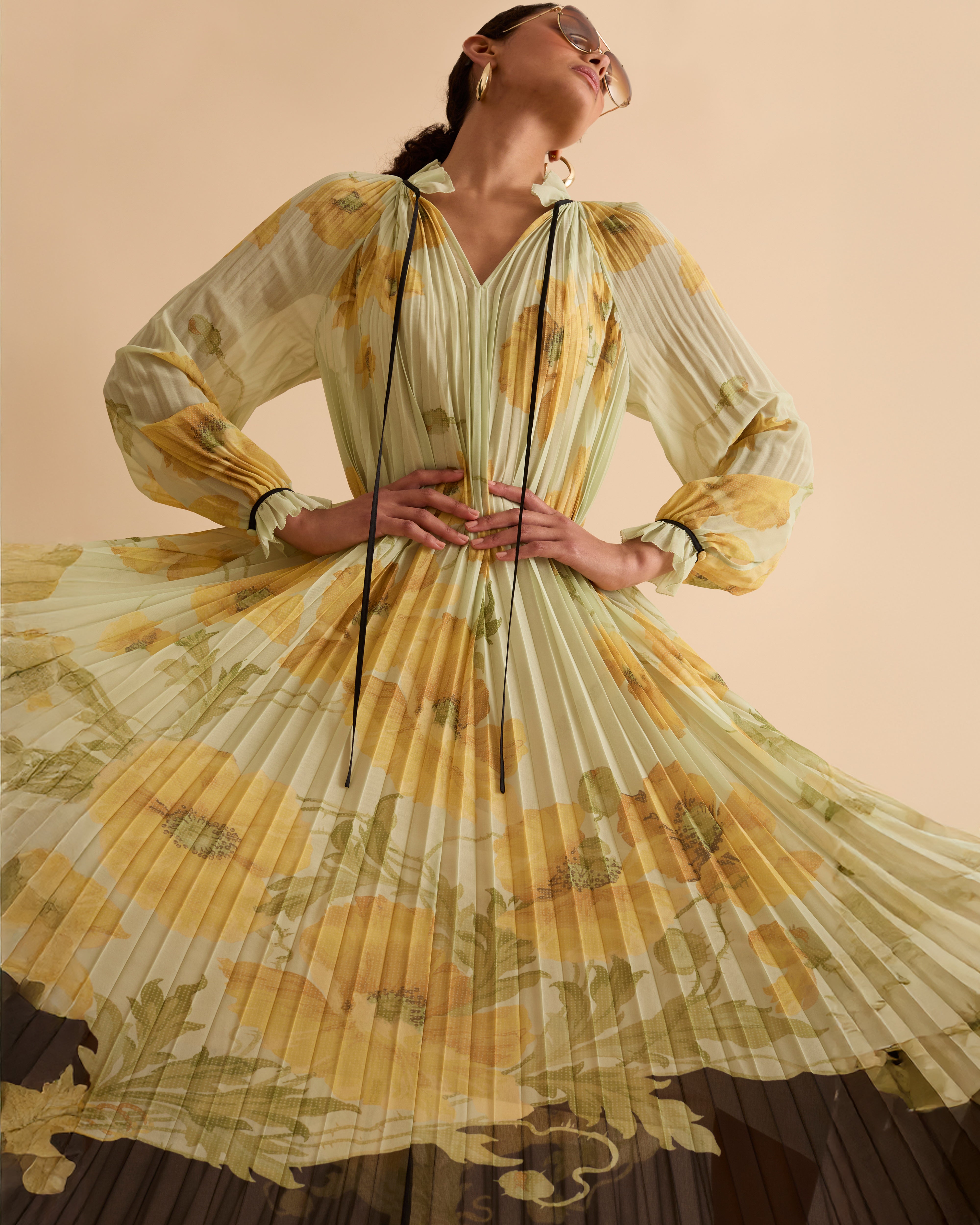 Model wearing Florere pleated floral maxi dress with yellow poppy print, sheer long sleeves, high neckline with ties and black hem, styled with gold hoop earrings and black open-toe boots against a beige background
