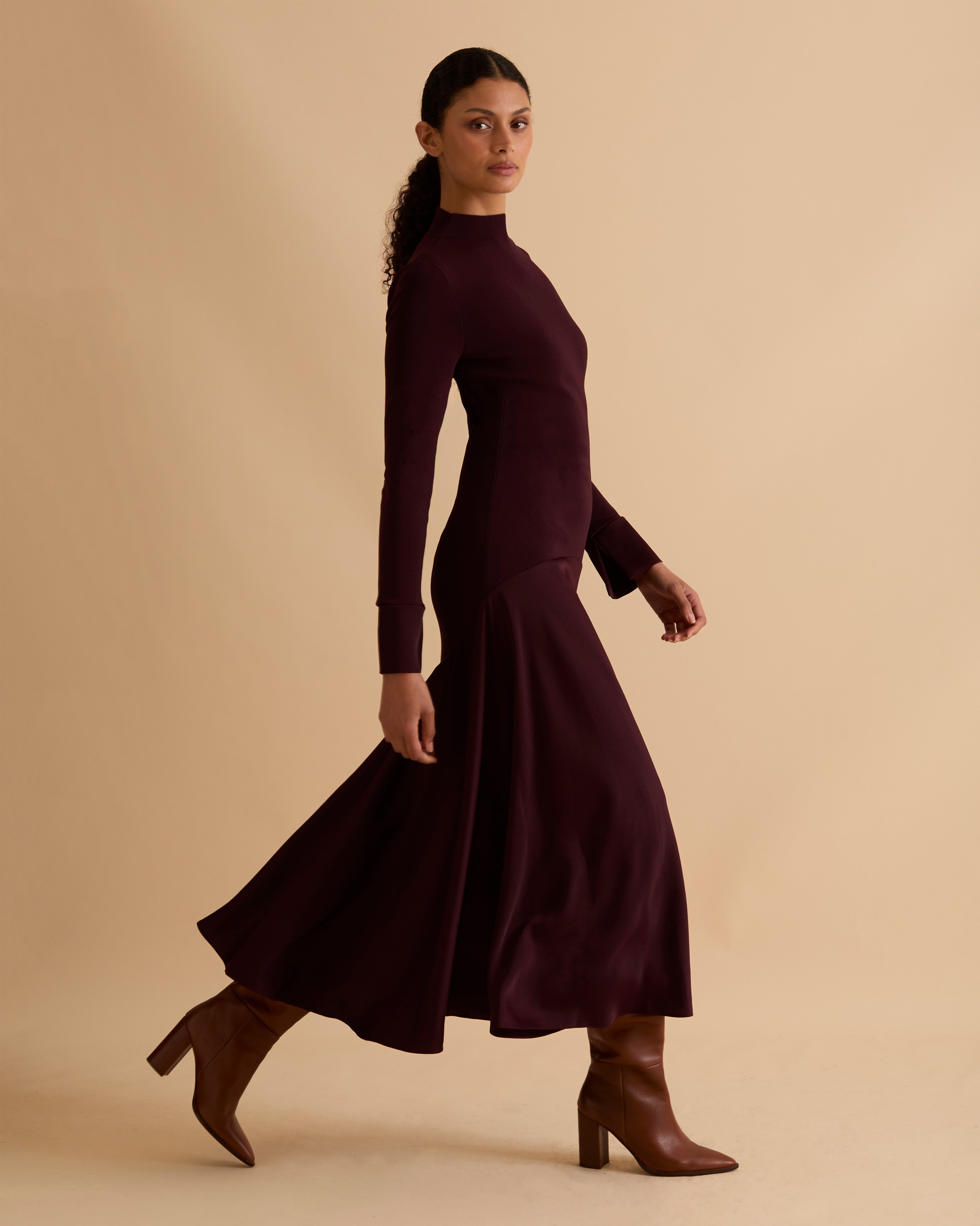 Model wearing Florere iconic signature Acacia dress. Knitted fabric for a fitted bodice, with dropped waist satin midi skirt with dramatic flare. Wearing tan knee high boots, against a beige neutral backdrop