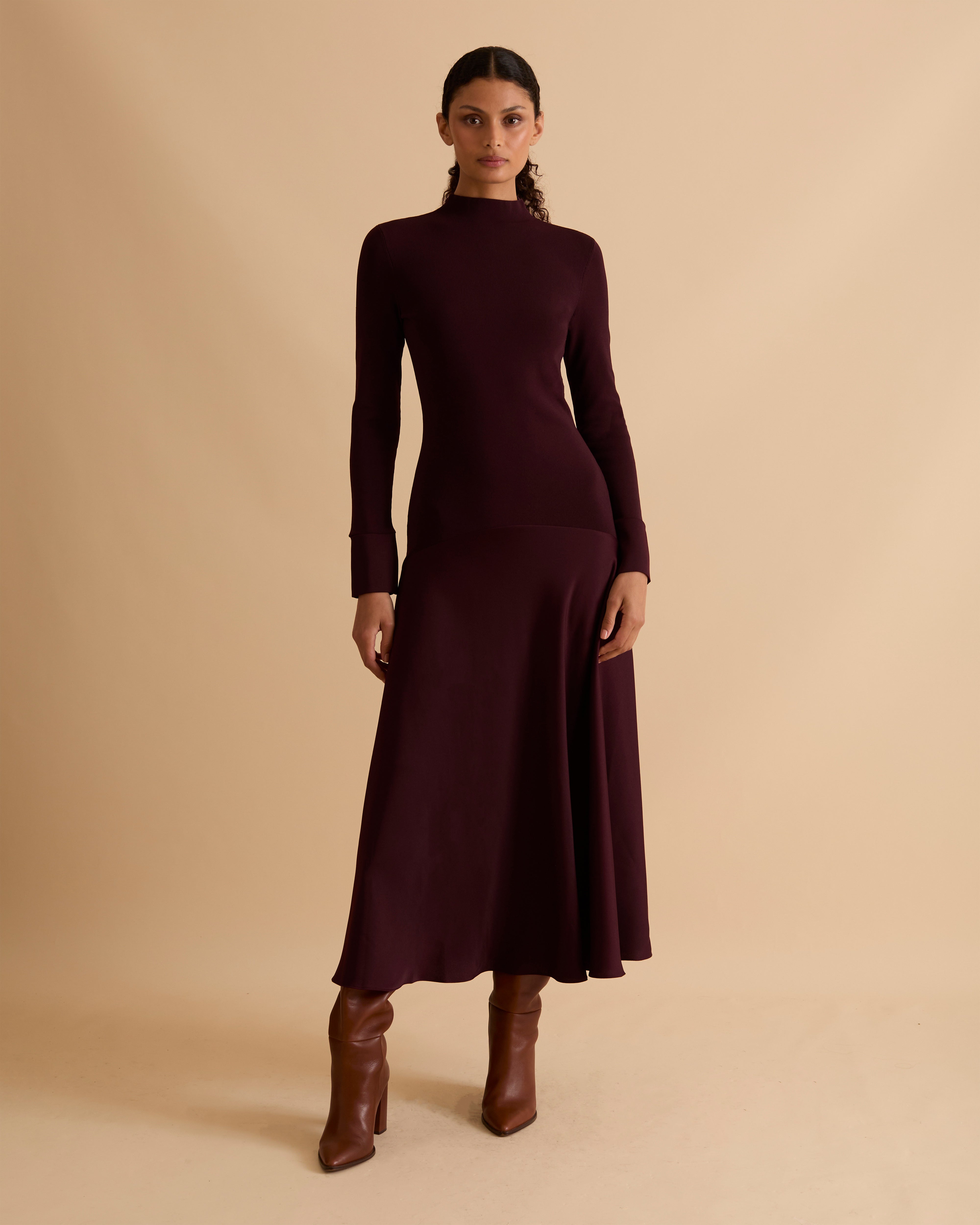 Model wearing Florere iconic signature Acacia dress. Knitted fabric for a fitted bodice, with dropped waist satin midi skirt with dramatic flare. Wearing tan knee high boots, against a beige neutral backdrop