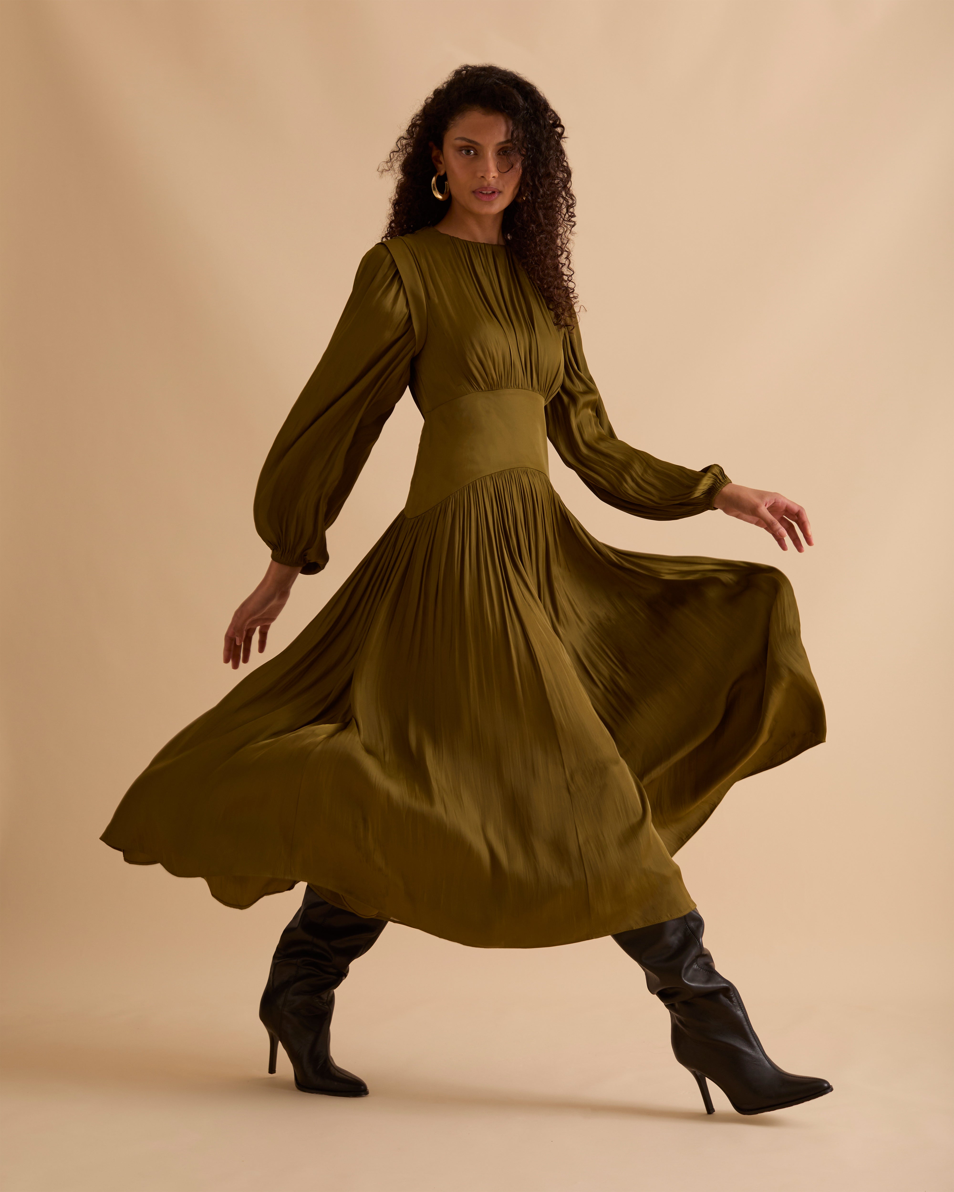 Model wearing Florere olive green maxi dress in billowing sating fabric, with high neck and long sleeves, flattering waist panel and gold zip at the back. Wearing black knee high boots and gold drop earrings against a beige, neutral backdrop
