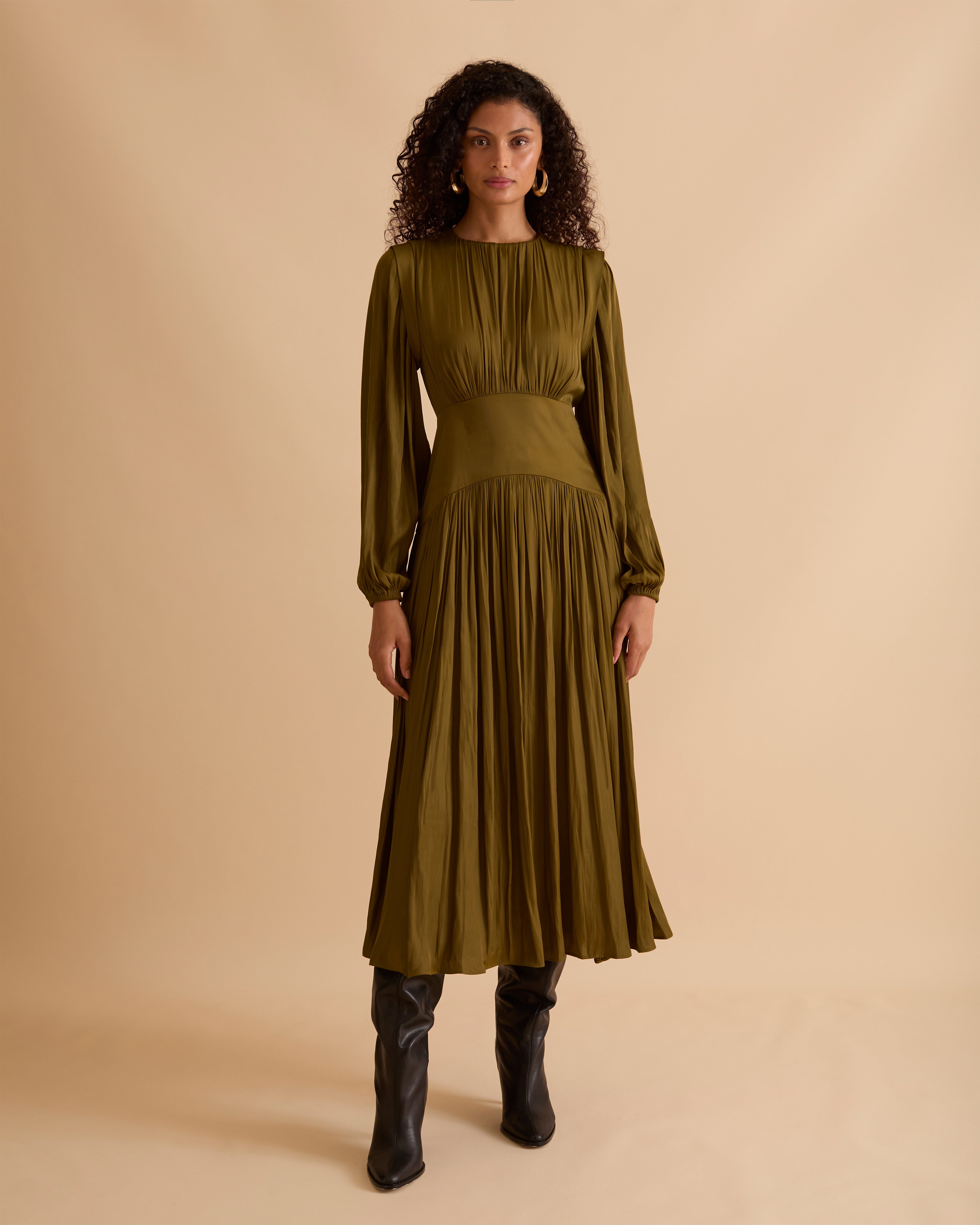 Model wearing Florere olive green maxi dress in billowing sating fabric, with high neck and long sleeves, flattering waist panel and gold zip at the back. Wearing black knee high boots and gold drop earrings against a beige, neutral backdrop