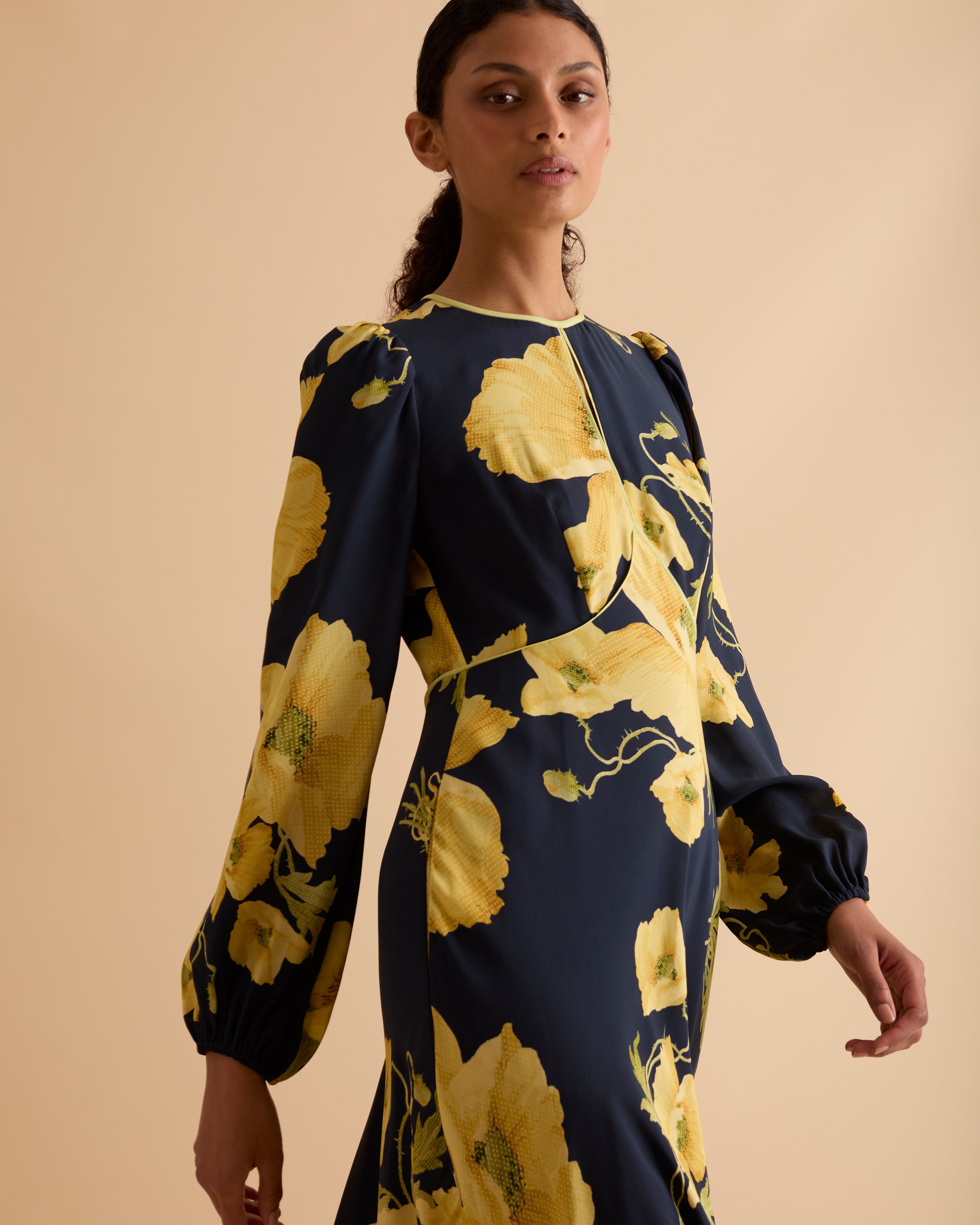 Model wearing Florere floral print occasionwear midi dress. Navy with yellow flowers, high neck dress, with long sleeves, fitted bodice, full skirt with pockets. Wearing tan knee high boots against a beige neutral backdrop