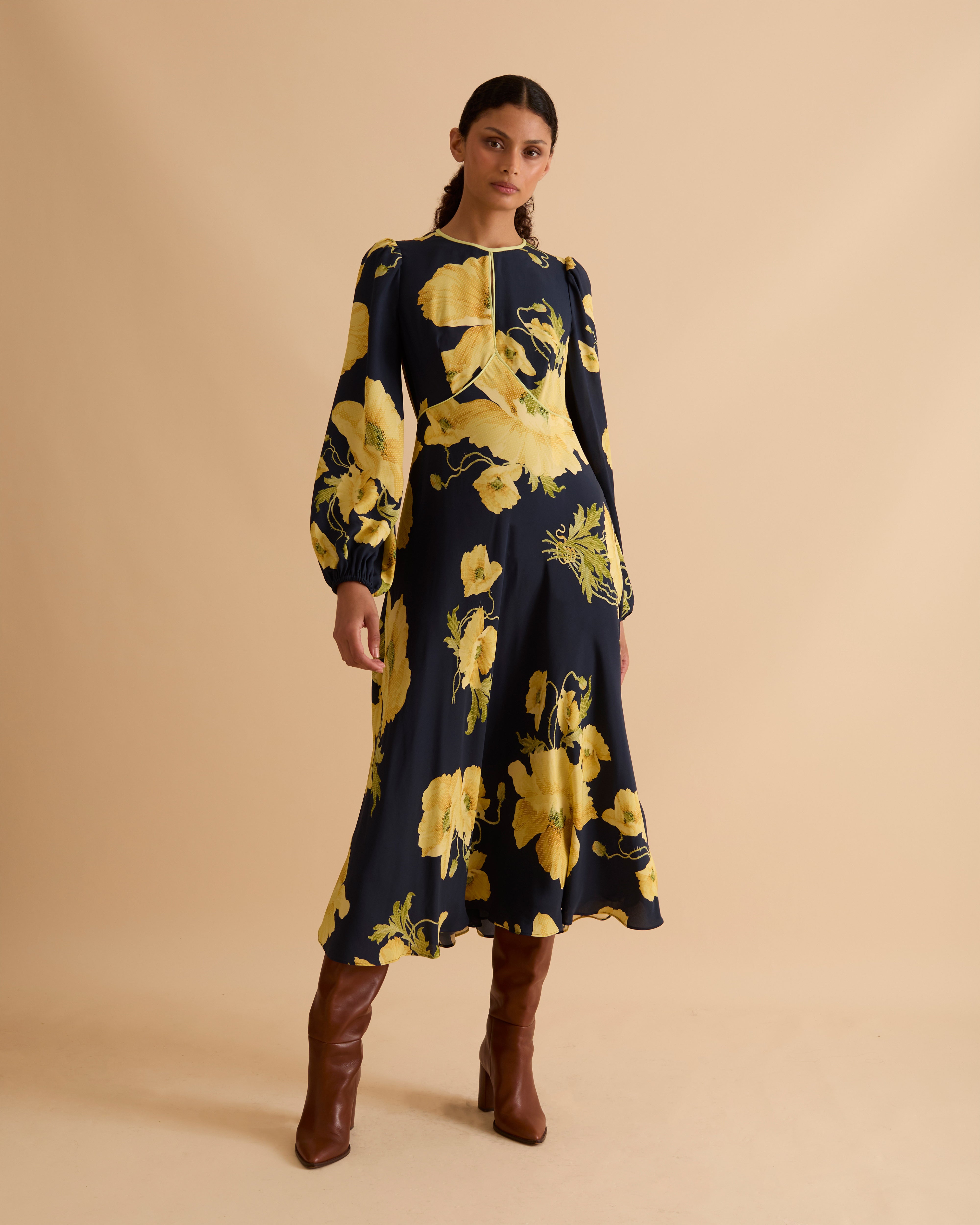 Model wearing Florere floral print occasionwear midi dress. Navy with yellow flowers, high neck dress, with long sleeves, fitted bodice, full skirt with pockets. Wearing tan knee high boots against a beige neutral backdrop