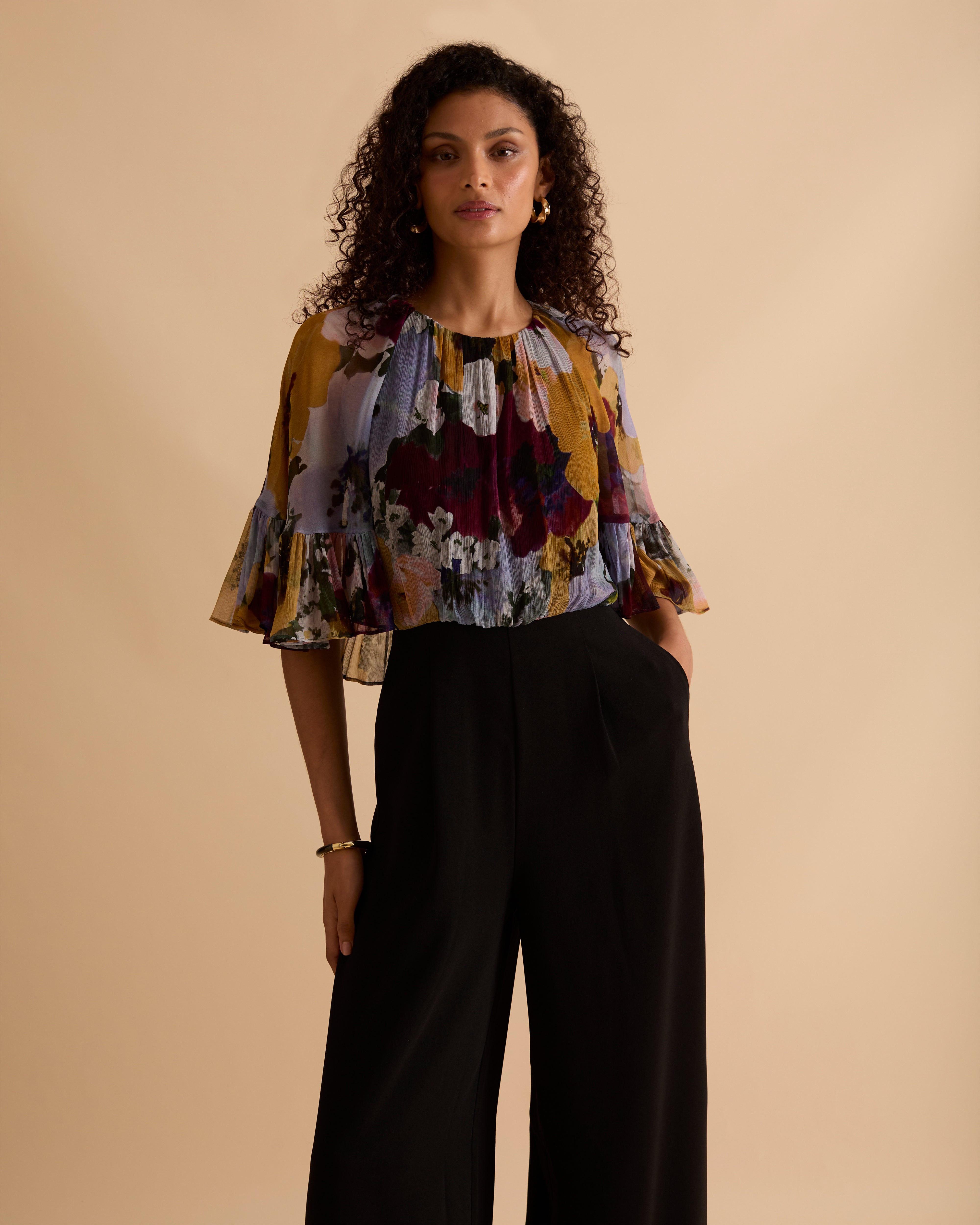 Model wearing Florere jumpsuit, with tailored black trouser and lightweight floral printed cape top, with high neck and covered arms. Against a beige neutral backdrop