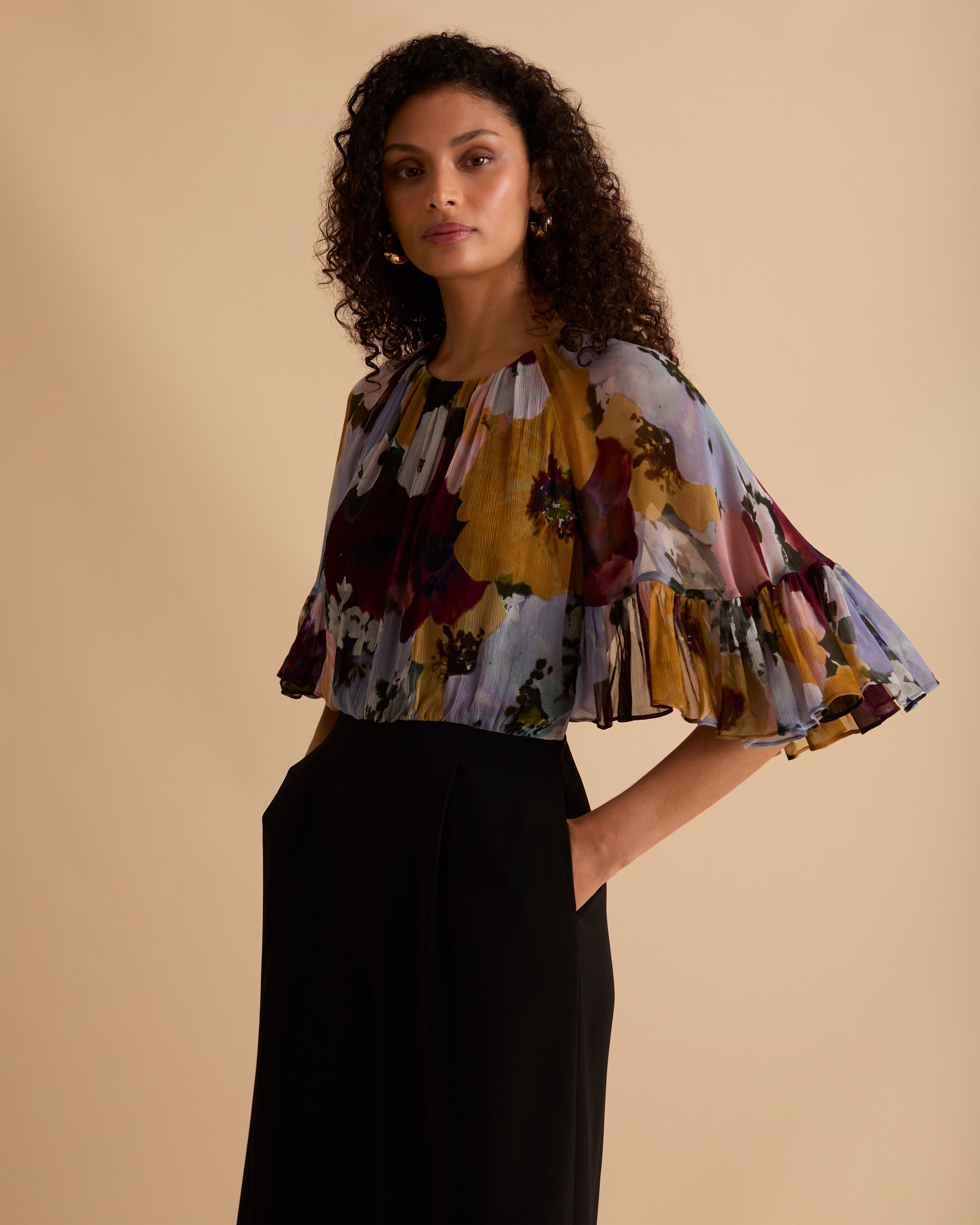 Model wearing Florere jumpsuit, with tailored black trouser and lightweight floral printed cape top, with high neck and covered arms. Against a beige neutral backdrop