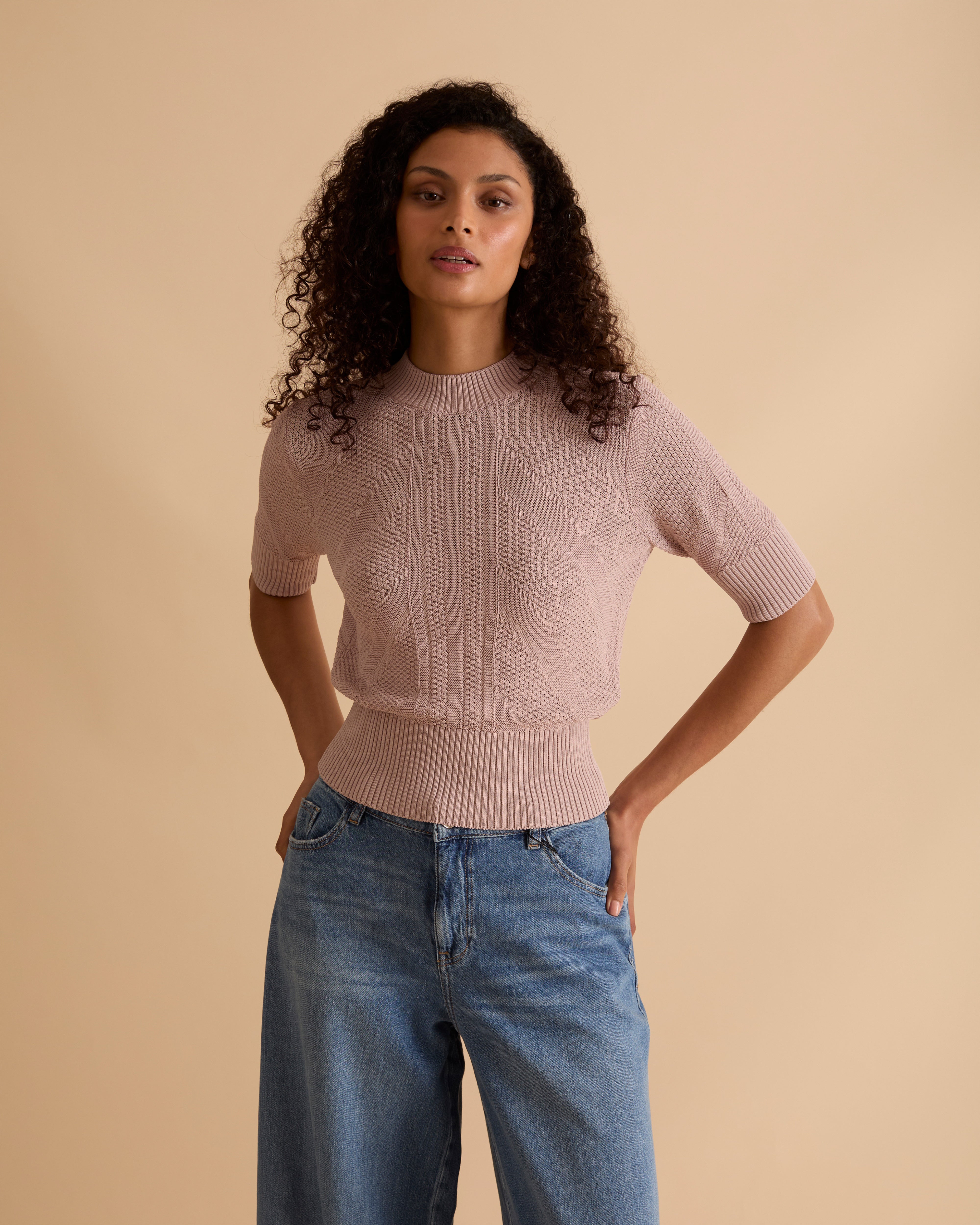 Model wearing Florere light pink, pointelle knit short sleeve lighweight jumper, with crew neck, worn with light blue wide legged jeans, against a beige neutral backdrop