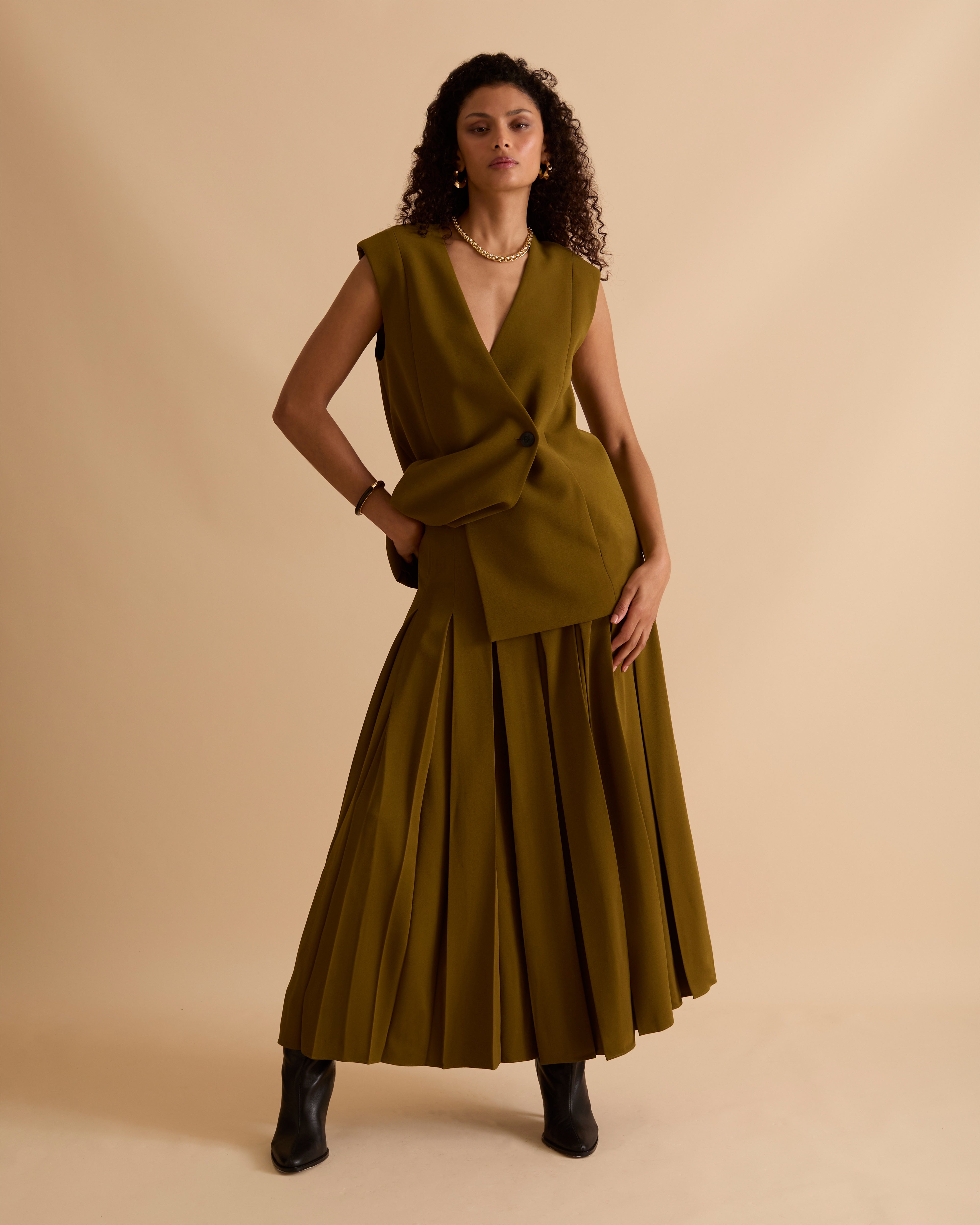 Model wearing Florere deep pleated midi skirt in olive green, wearing black boots and matching sleeveless waistcoat with gold chunky necklace on a beige neutral backdrop