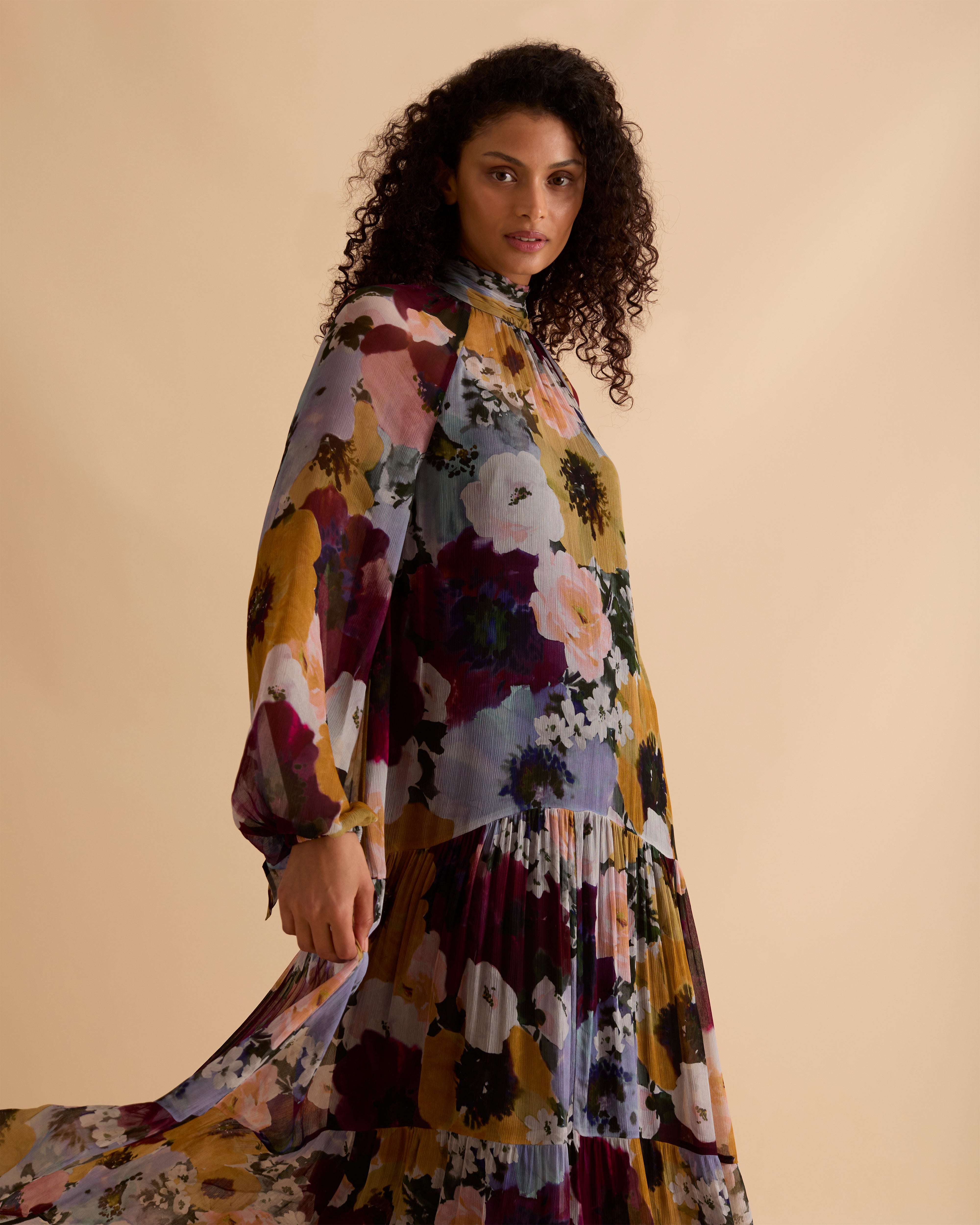 Model wearing Florere floral print tiered maxi dress, with high neckline, long sleeves, and neck tie to the back, styled with burgundy slouch boots, against a neutral beige background