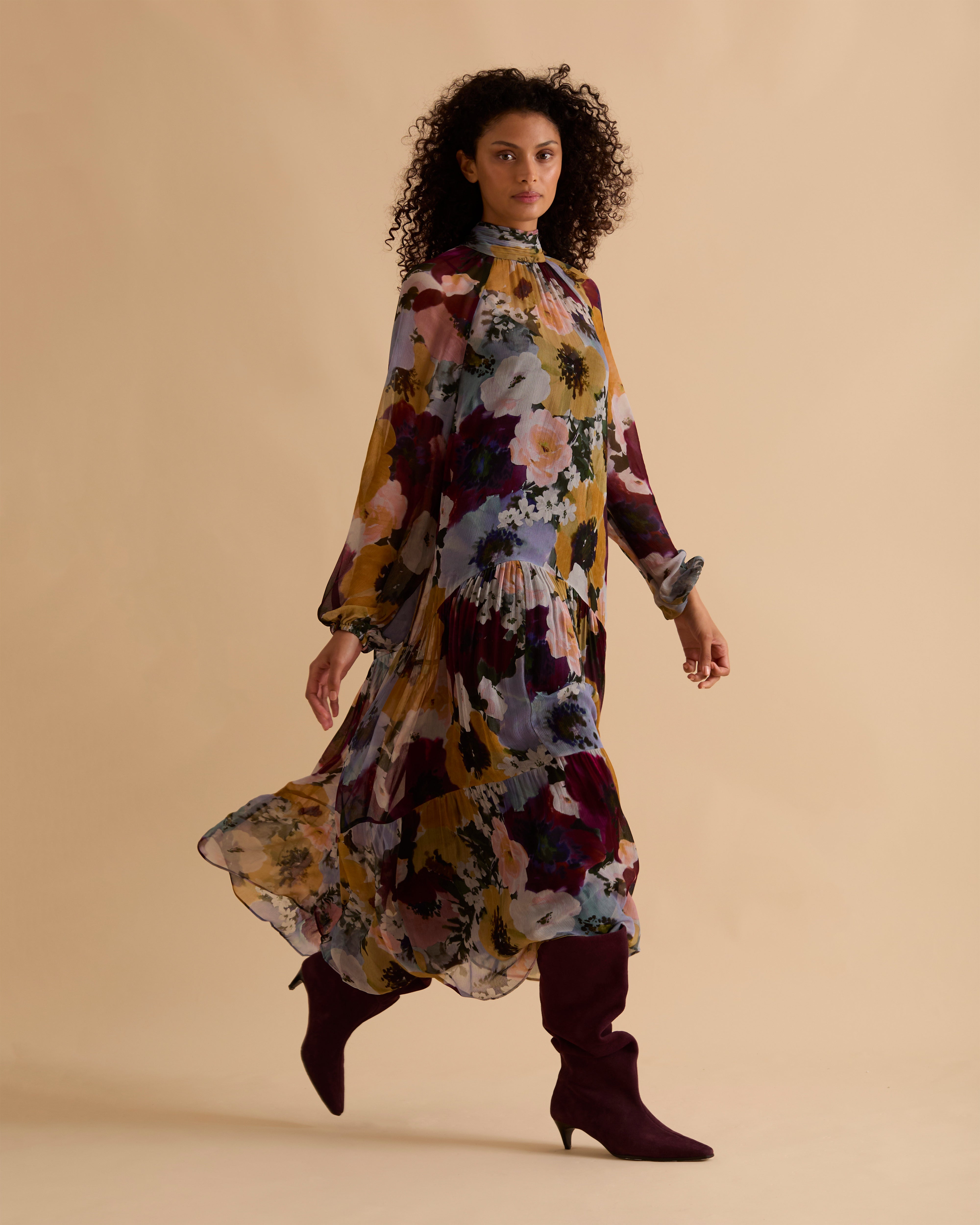 Model wearing Florere floral print tiered maxi dress, with high neckline, long sleeves, and neck tie to the back, styled with burgundy slouch boots, against a neutral beige background
