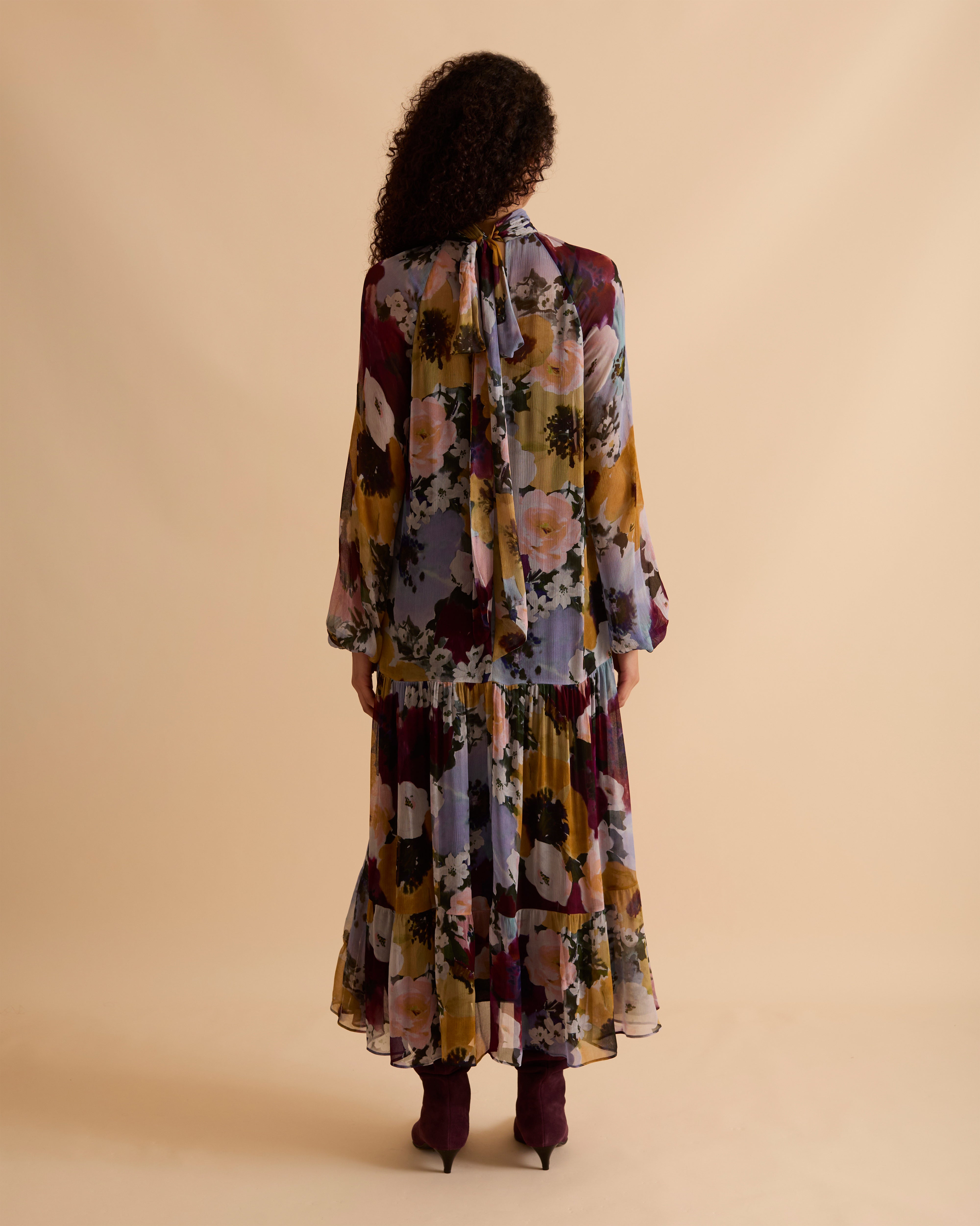 Model wearing Florere floral print tiered maxi dress, with high neckline, long sleeves, and neck tie to the back, styled with burgundy slouch boots, against a neutral beige background