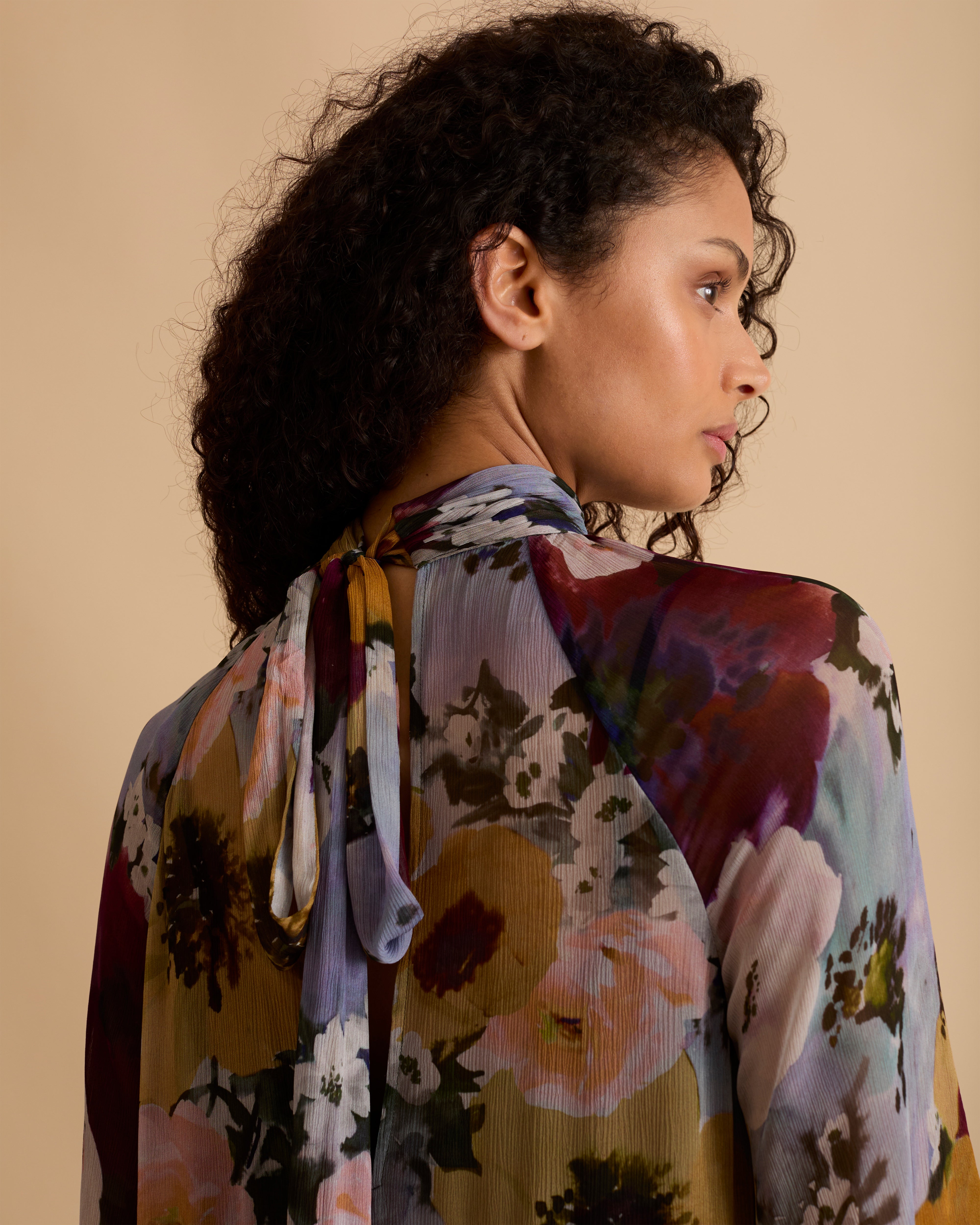 Model wearing Florere floral print tiered maxi dress, with high neckline, long sleeves, and neck tie to the back, styled with burgundy slouch boots, against a neutral beige background