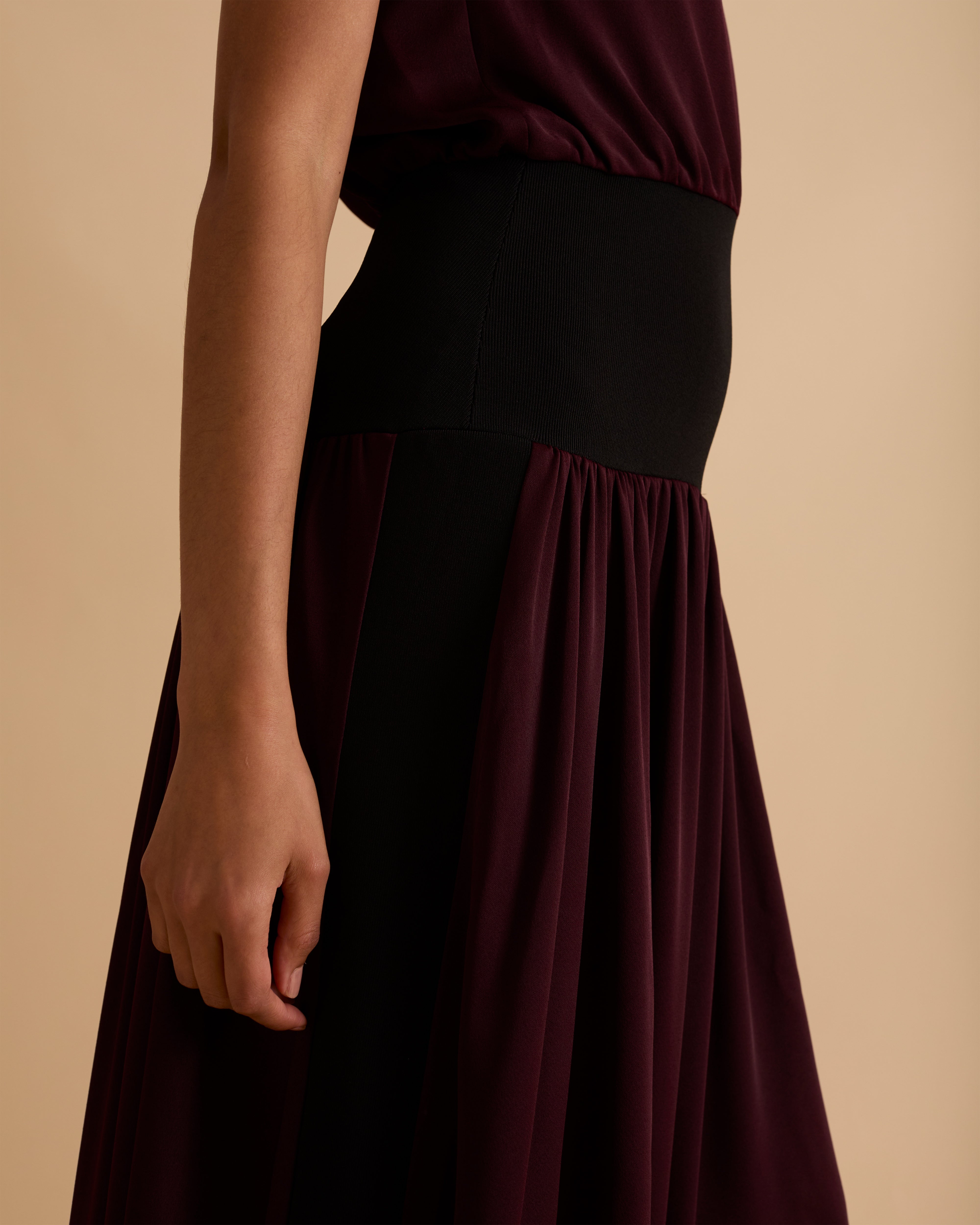 Model wearing Florere sleeveless burgundy midi dress with high neckline, black waistband detail, and flowy A-line silhouette, styled with gold hoop earrings and black open-toe heels, standing against a beige background