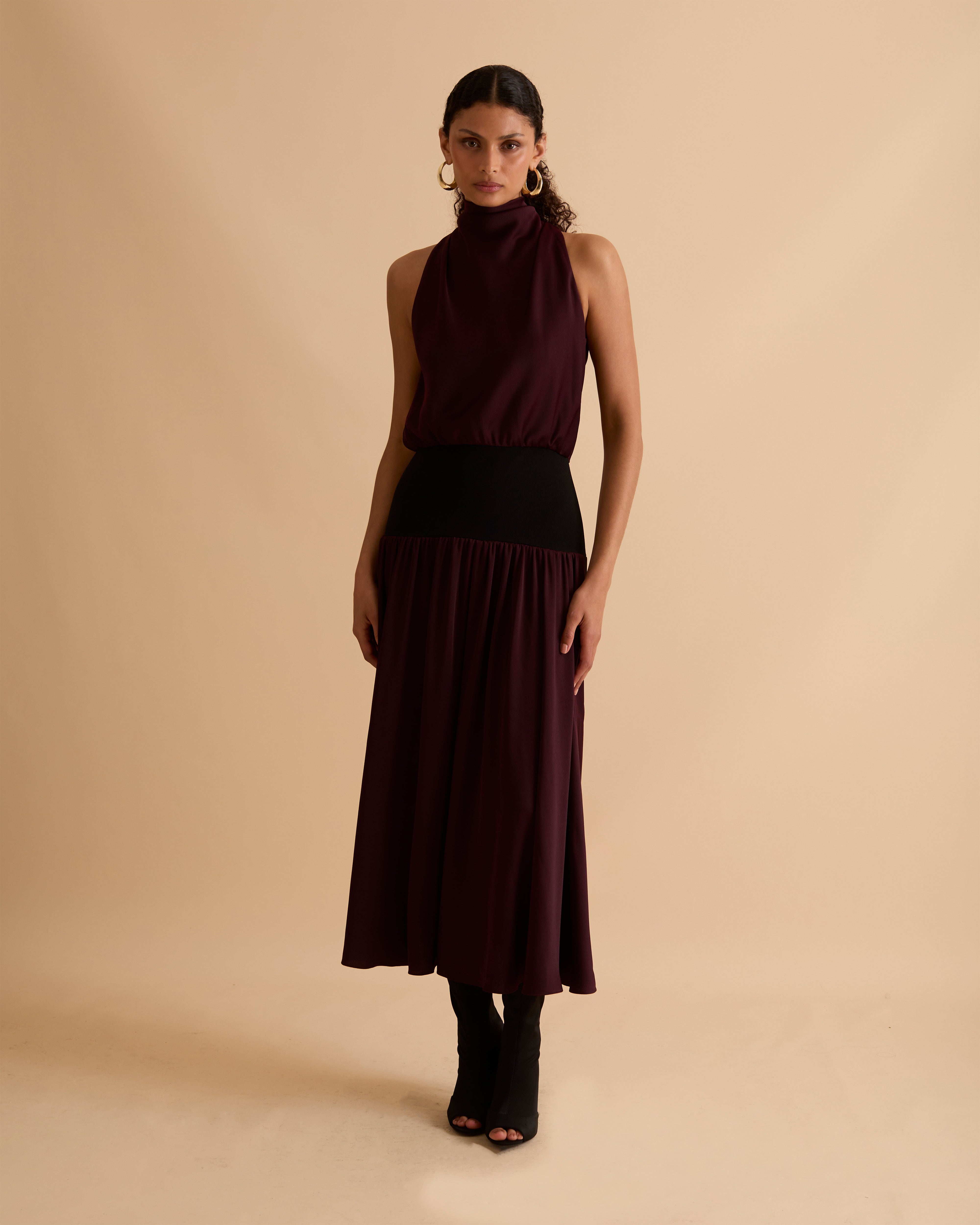 Model wearing Florere sleeveless burgundy midi dress with high neckline, black waistband detail, and flowy A-line silhouette, styled with gold hoop earrings and black open-toe heels, standing against a beige background
