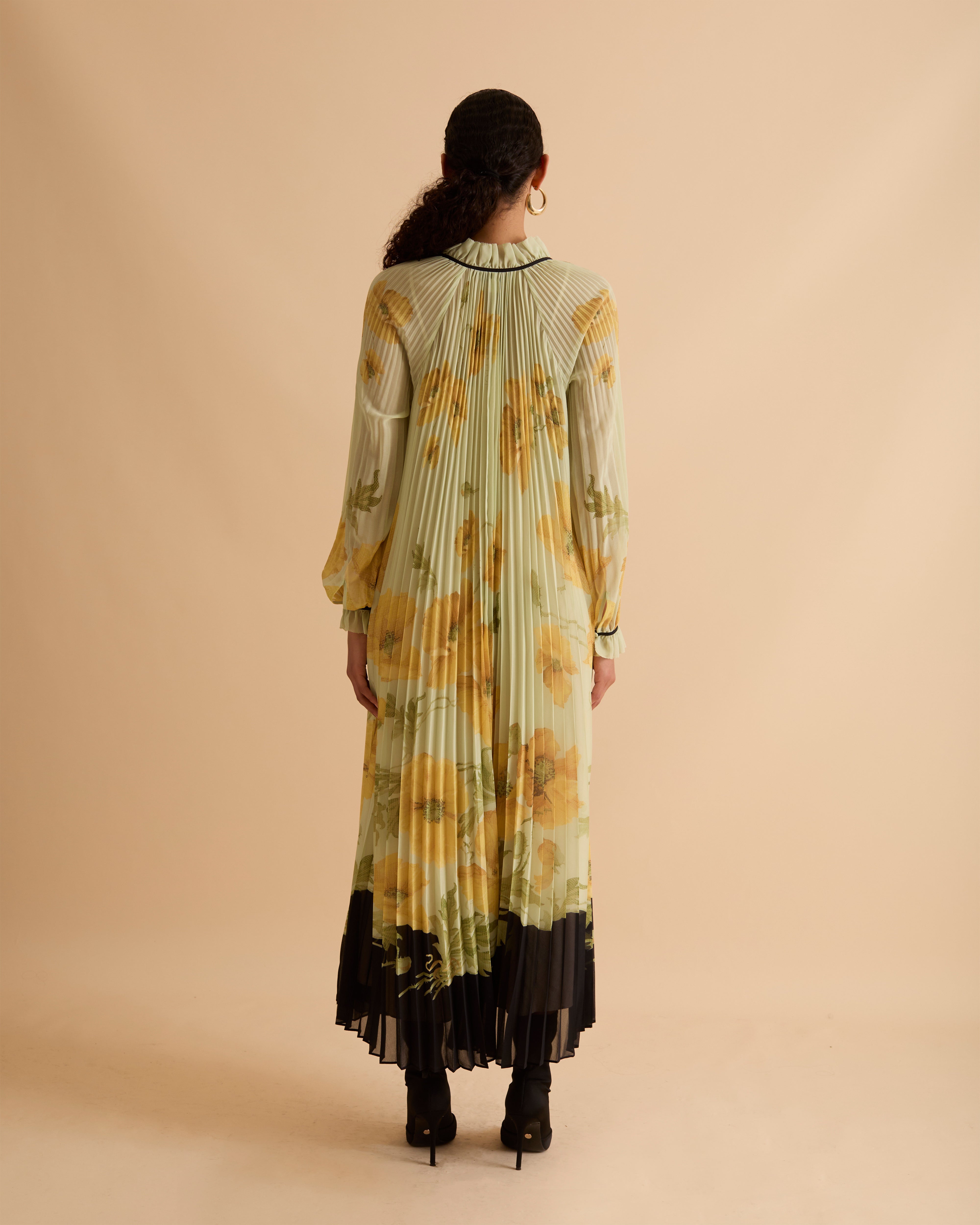 Model wearing Florere pleated floral maxi dress with yellow poppy print, sheer long sleeves, high neckline with ties and black hem, styled with gold hoop earrings and black open-toe boots against a beige background