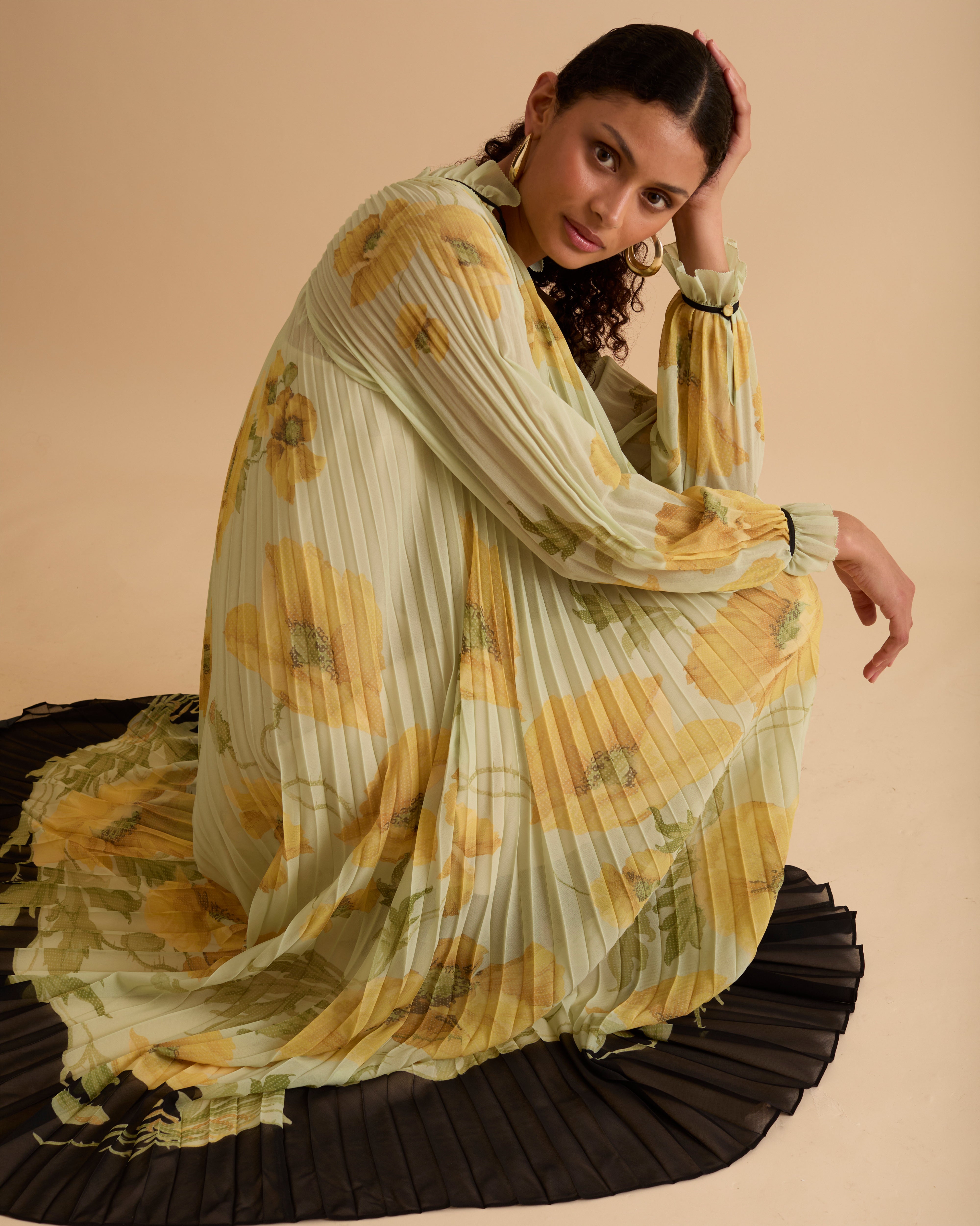 Model wearing Florere pleated floral maxi dress with yellow poppy print, sheer long sleeves, high neckline with ties and black hem, styled with gold hoop earrings and black open-toe boots against a beige background