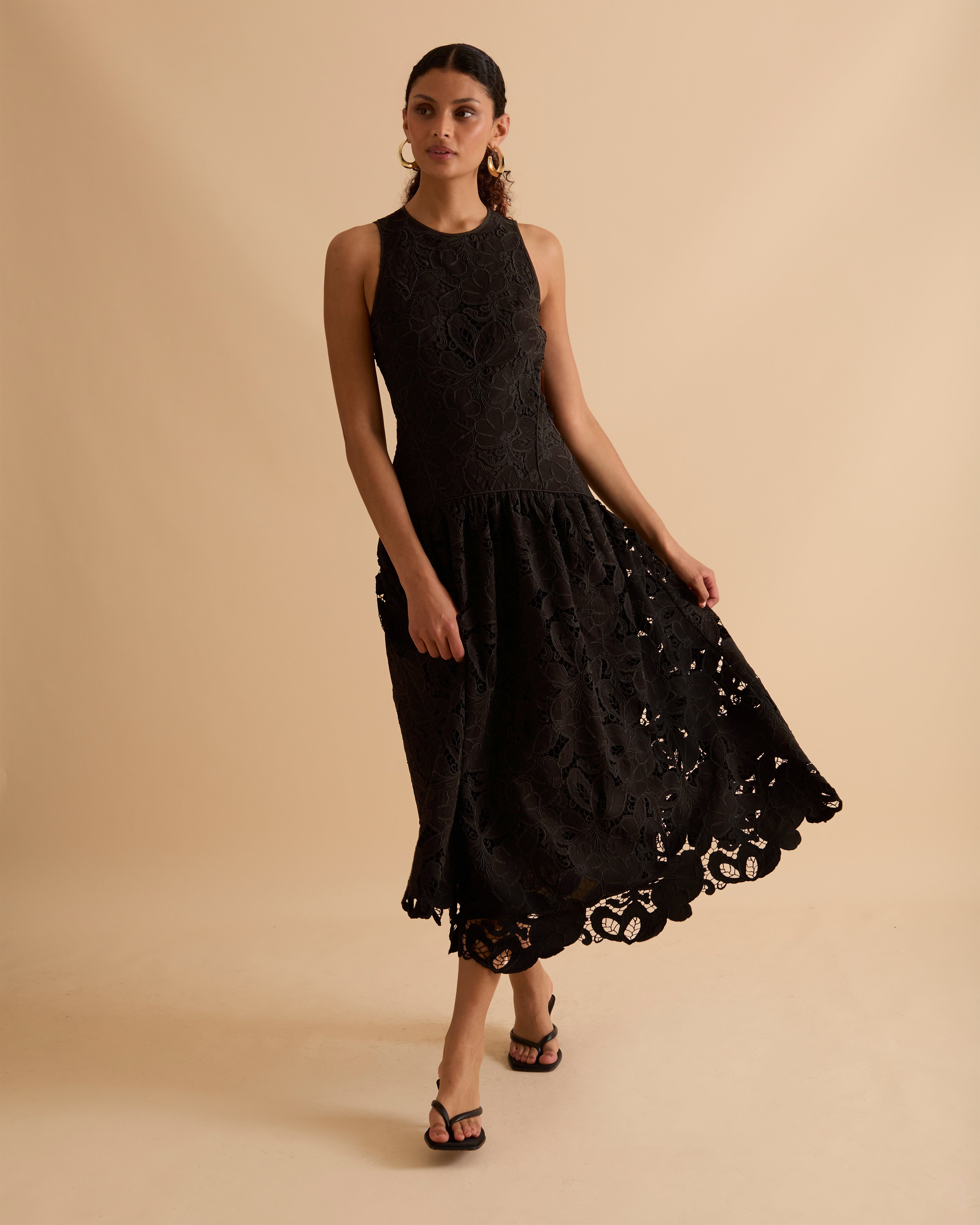 Model wearing Florere black lace midi dress, with scallop hem, panelled fitted bodice and fit and flare skirt. Wearing gold hoop statement earrings, against a beige backdrop