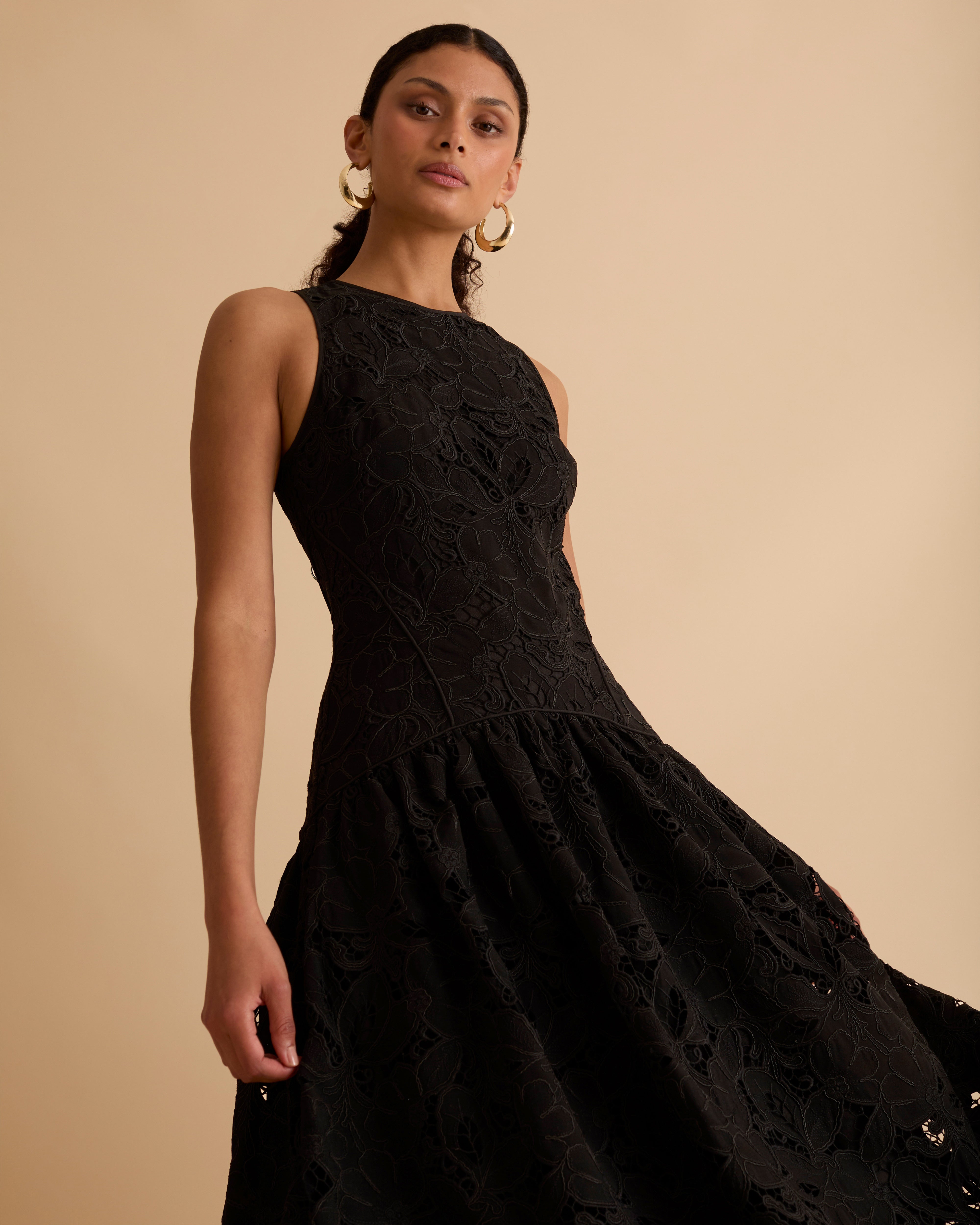 Model wearing Florere black lace midi dress, with scallop hem, panelled fitted bodice and fit and flare skirt. Wearing gold hoop statement earrings, against a beige backdrop