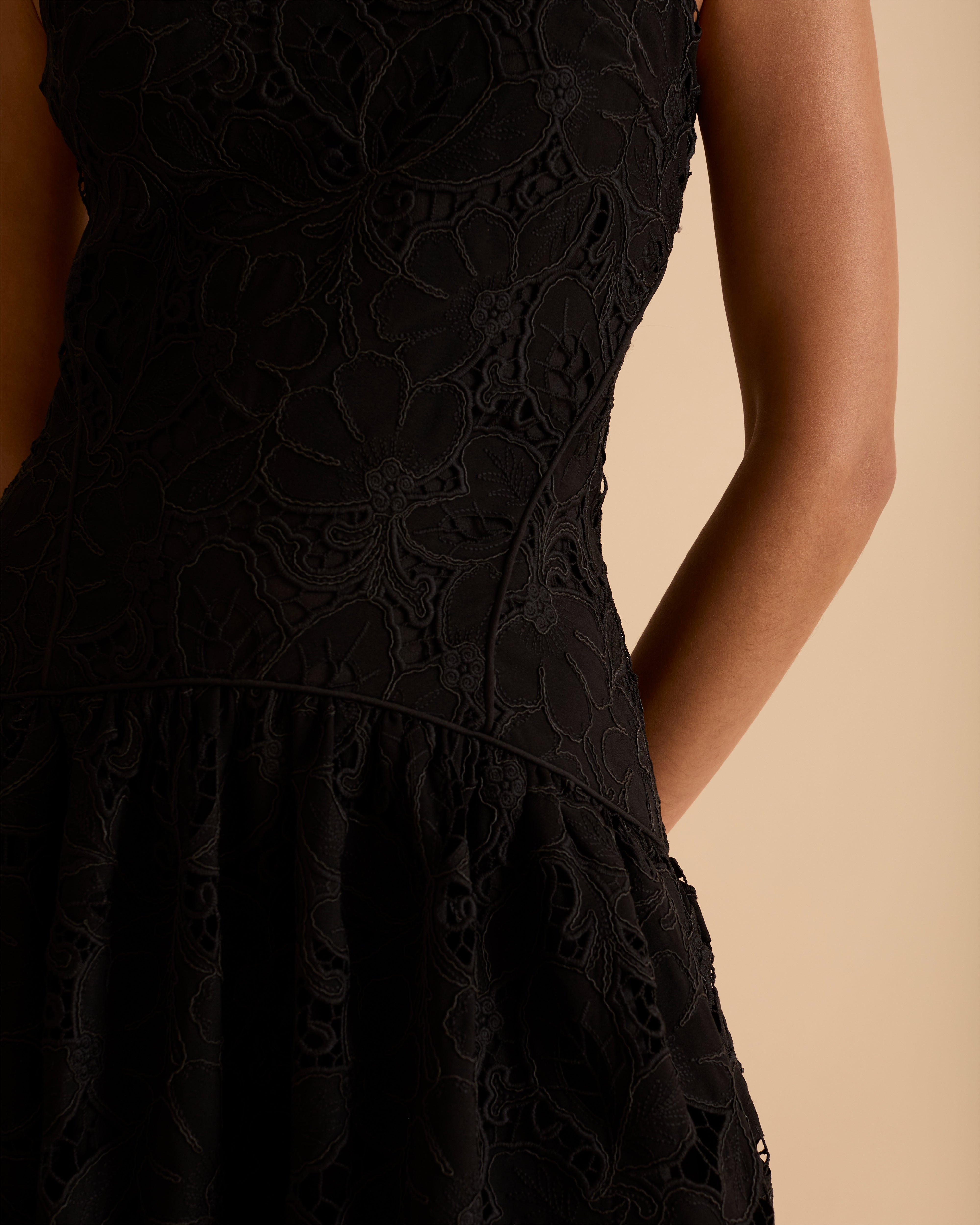 Model wearing Florere black lace midi dress, with scallop hem, panelled fitted bodice and fit and flare skirt. Wearing gold hoop statement earrings, against a beige backdrop