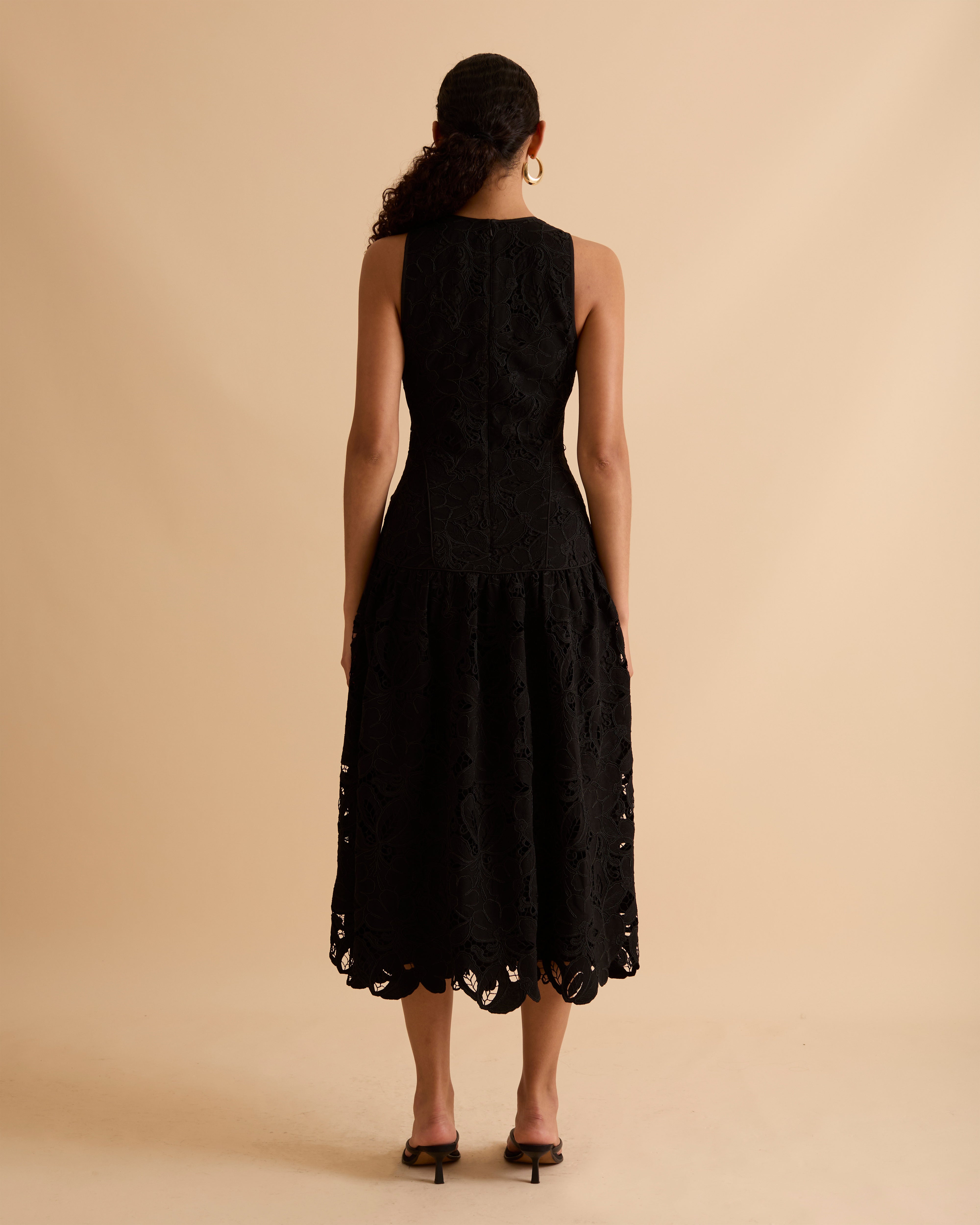 Model wearing Florere black lace midi dress, with scallop hem, panelled fitted bodice and fit and flare skirt. Wearing gold hoop statement earrings, against a beige backdrop