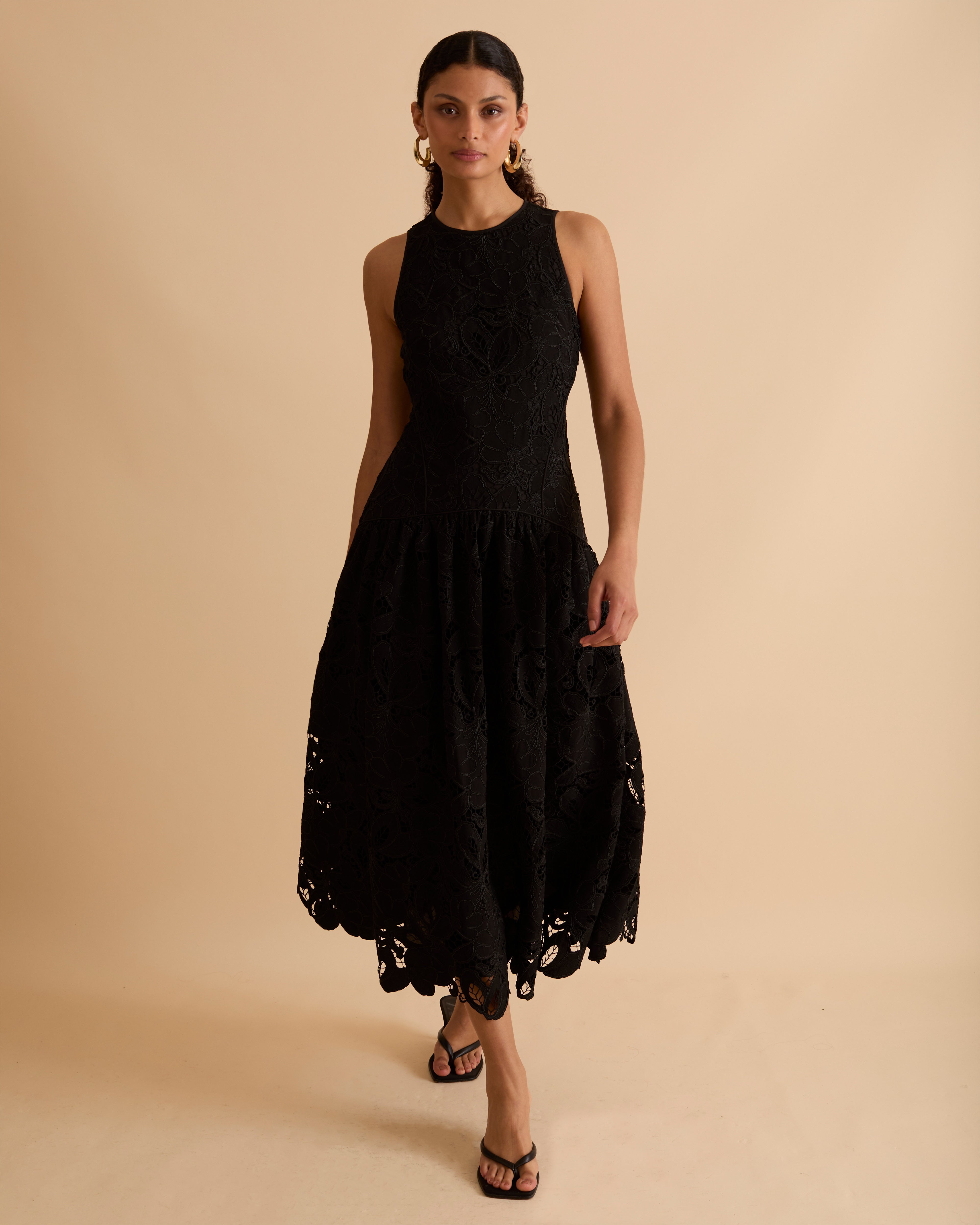 Model wearing Florere black lace midi dress, with scallop hem, panelled fitted bodice and fit and flare skirt. Wearing gold hoop statement earrings, against a beige backdrop