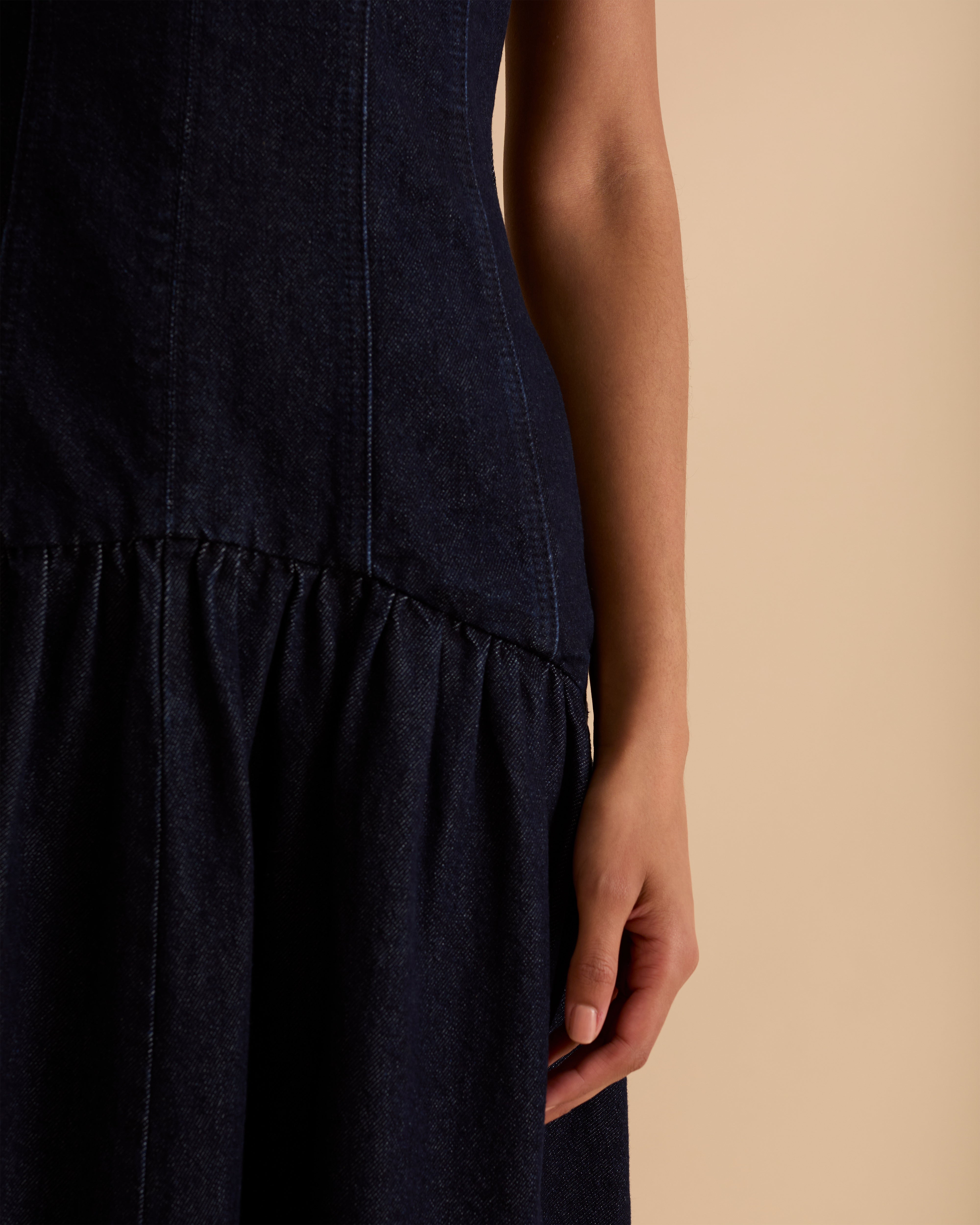 Model wearing Florere dark denim midi dress, with seamed fitted bodice and zip to the back, dropped waist skirt with dramatic volume skirt. Wearing tan knee high boots, against a beige neutral backdrop
