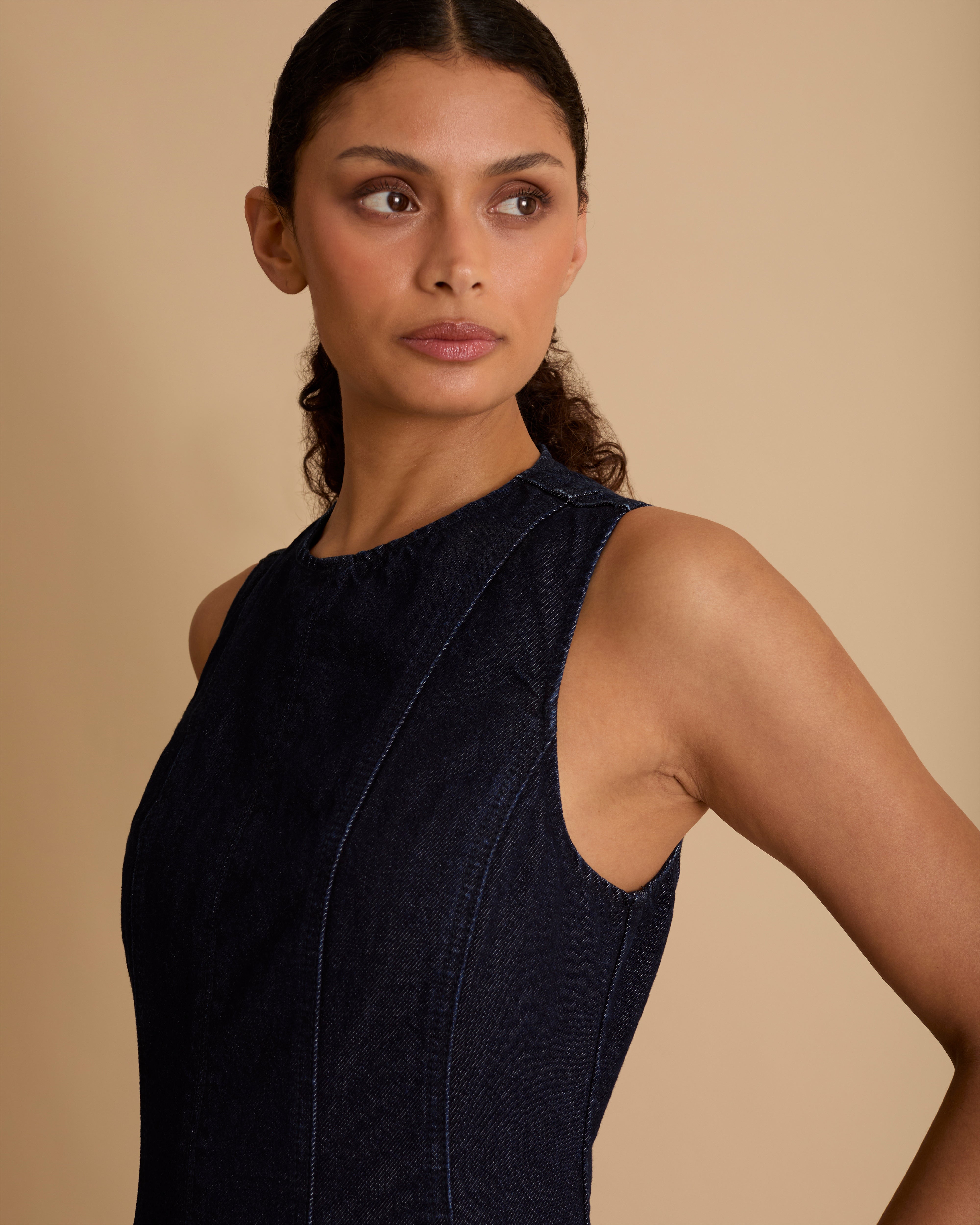 Model wearing Florere dark denim midi dress, with seamed fitted bodice and zip to the back, dropped waist skirt with dramatic volume skirt. Wearing tan knee high boots, against a beige neutral backdrop