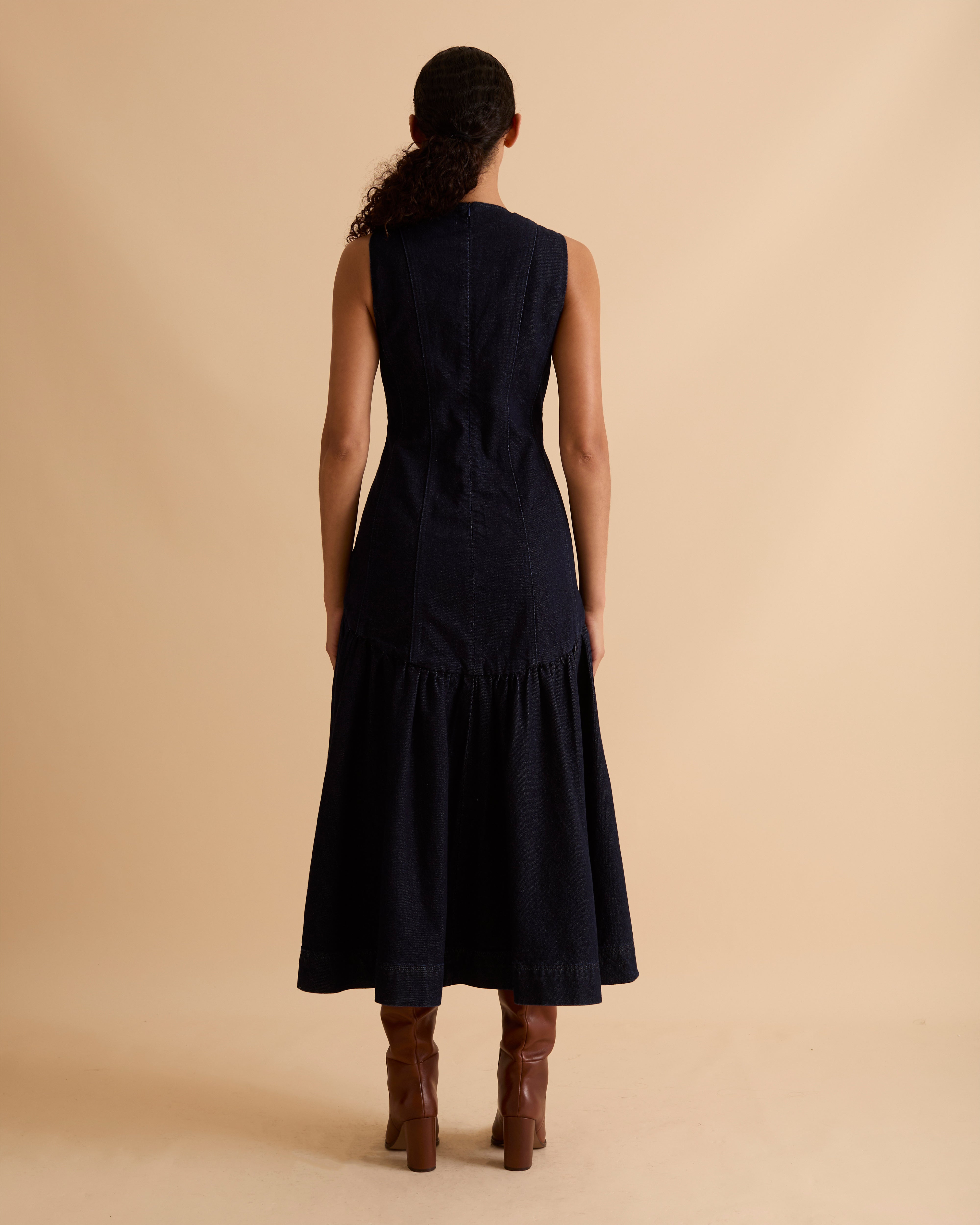 Model wearing Florere dark denim midi dress, with seamed fitted bodice and zip to the back, dropped waist skirt with dramatic volume skirt. Wearing tan knee high boots, against a beige neutral backdrop