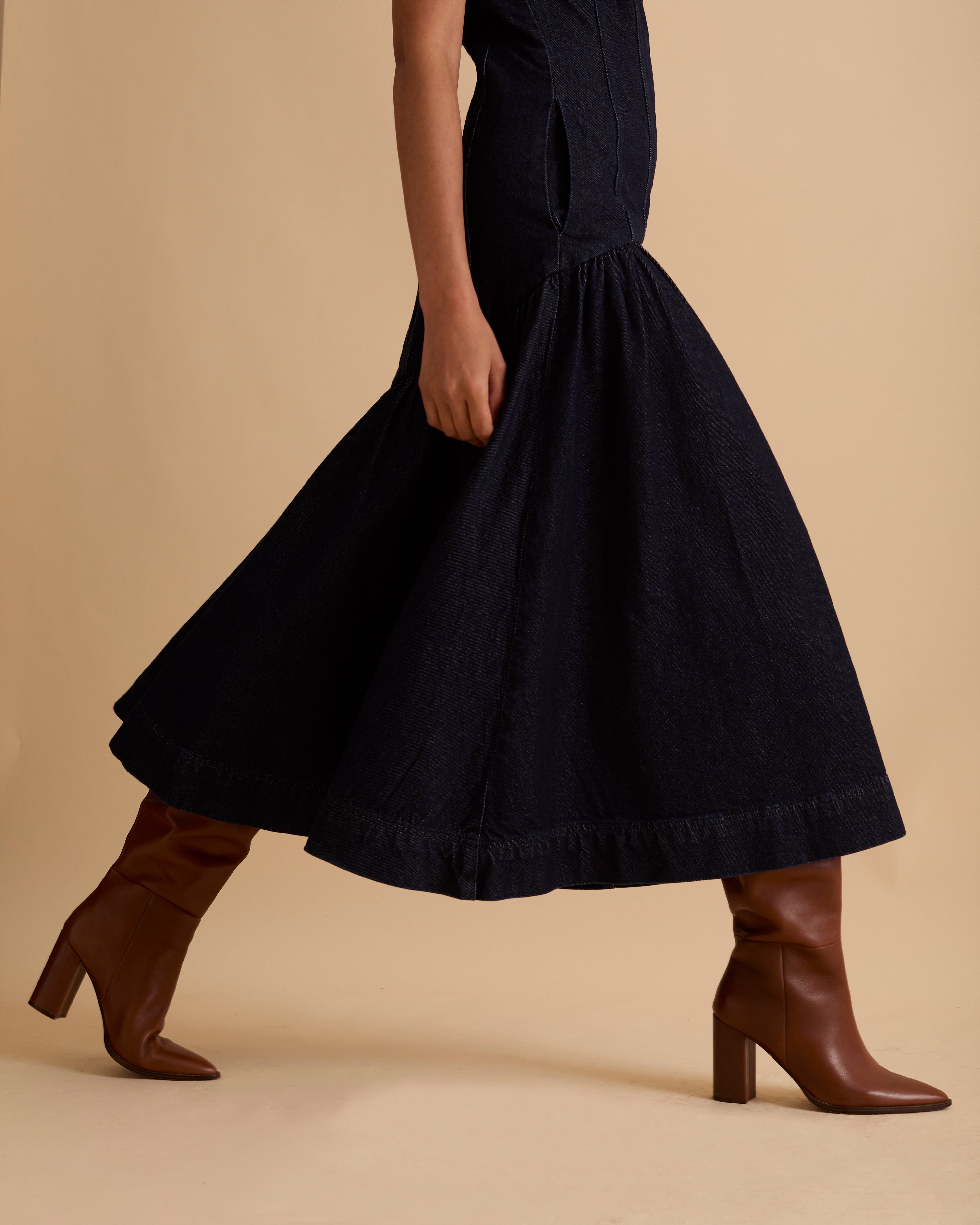 Model wearing Florere dark denim midi dress, with seamed fitted bodice and zip to the back, dropped waist skirt with dramatic volume skirt. Wearing tan knee high boots, against a beige neutral backdrop