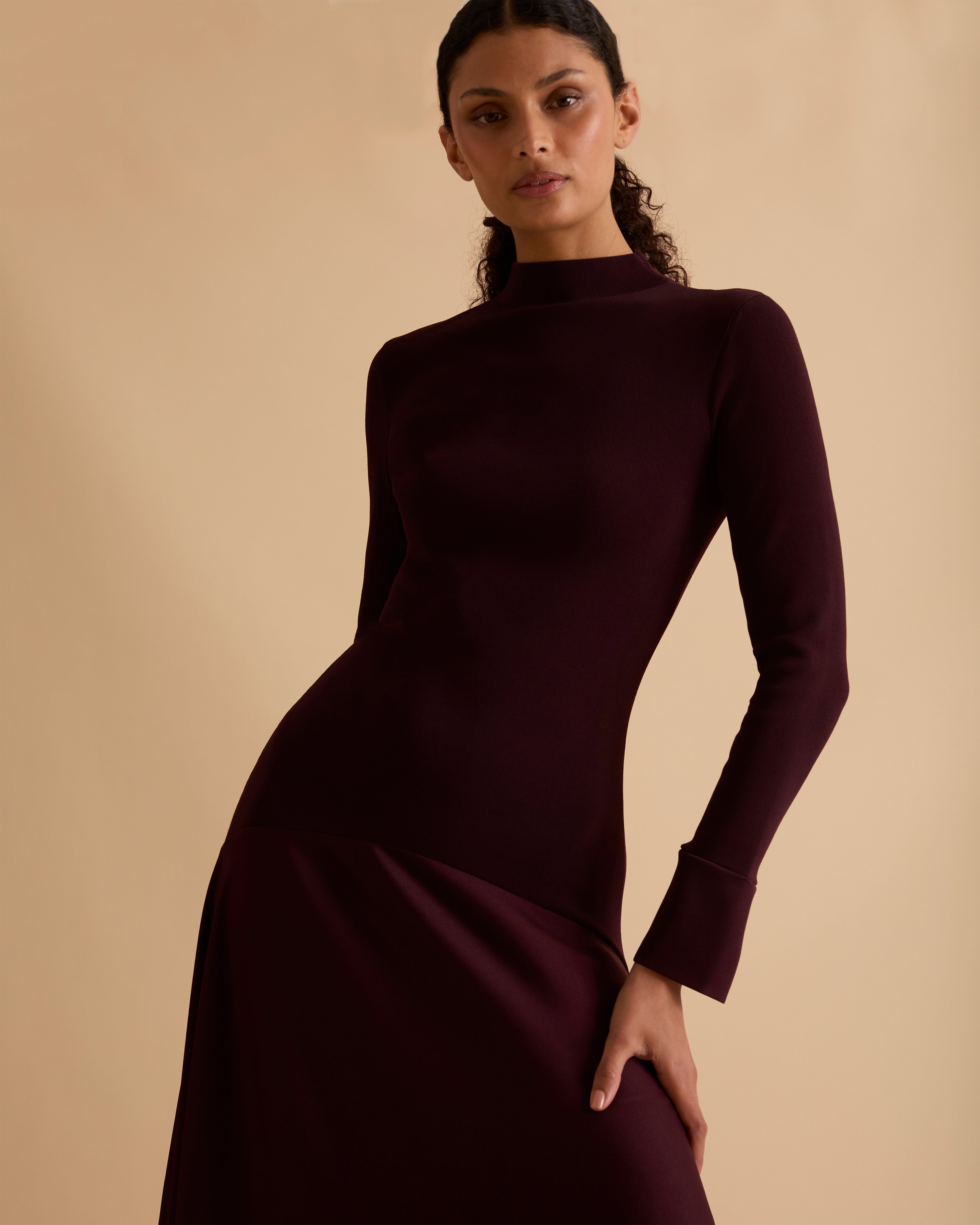 Model wearing Florere iconic signature Acacia dress. Knitted fabric for a fitted bodice, with dropped waist satin midi skirt with dramatic flare. Wearing tan knee high boots, against a beige neutral backdrop