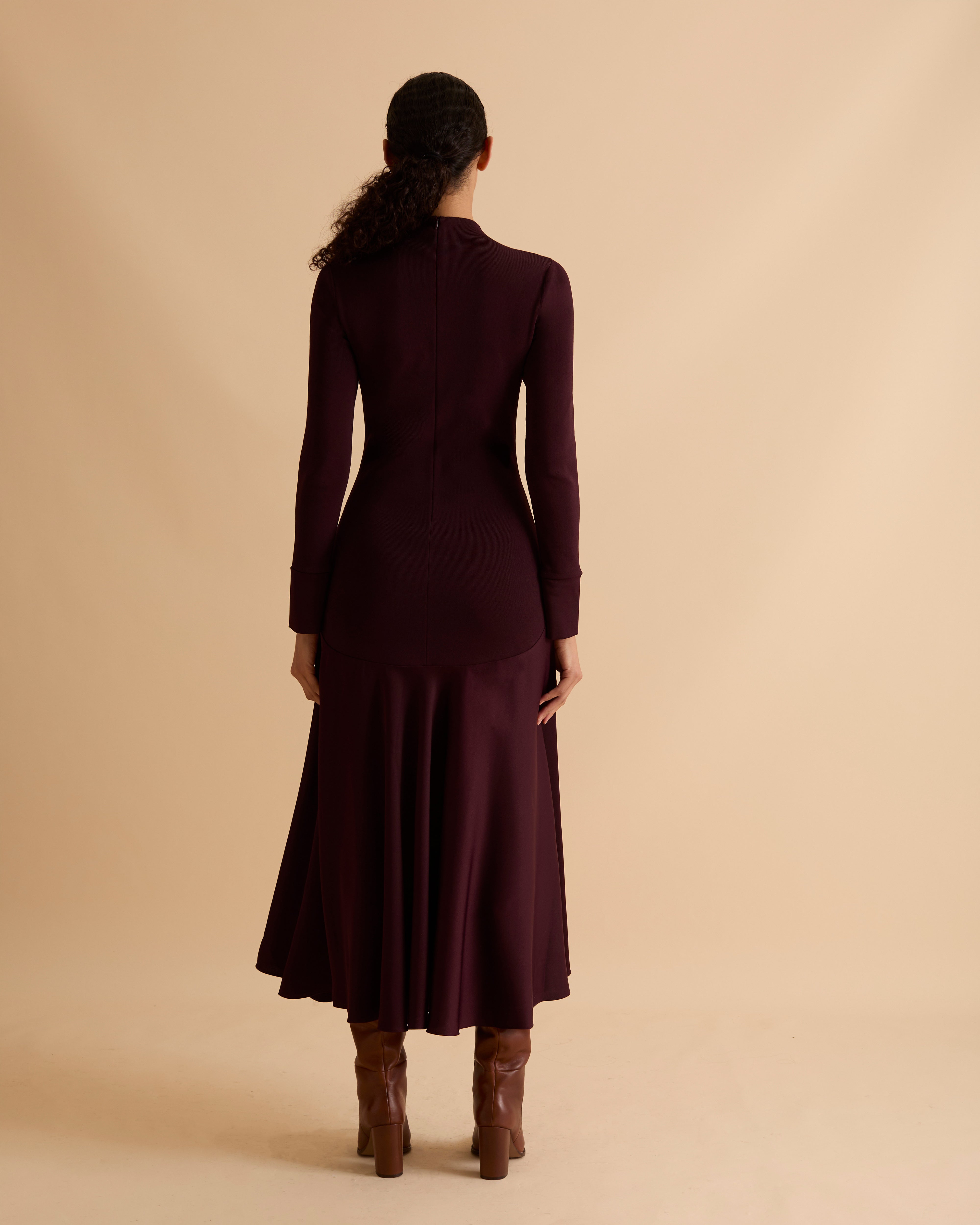 Model wearing Florere iconic signature Acacia dress. Knitted fabric for a fitted bodice, with dropped waist satin midi skirt with dramatic flare. Wearing tan knee high boots, against a beige neutral backdrop