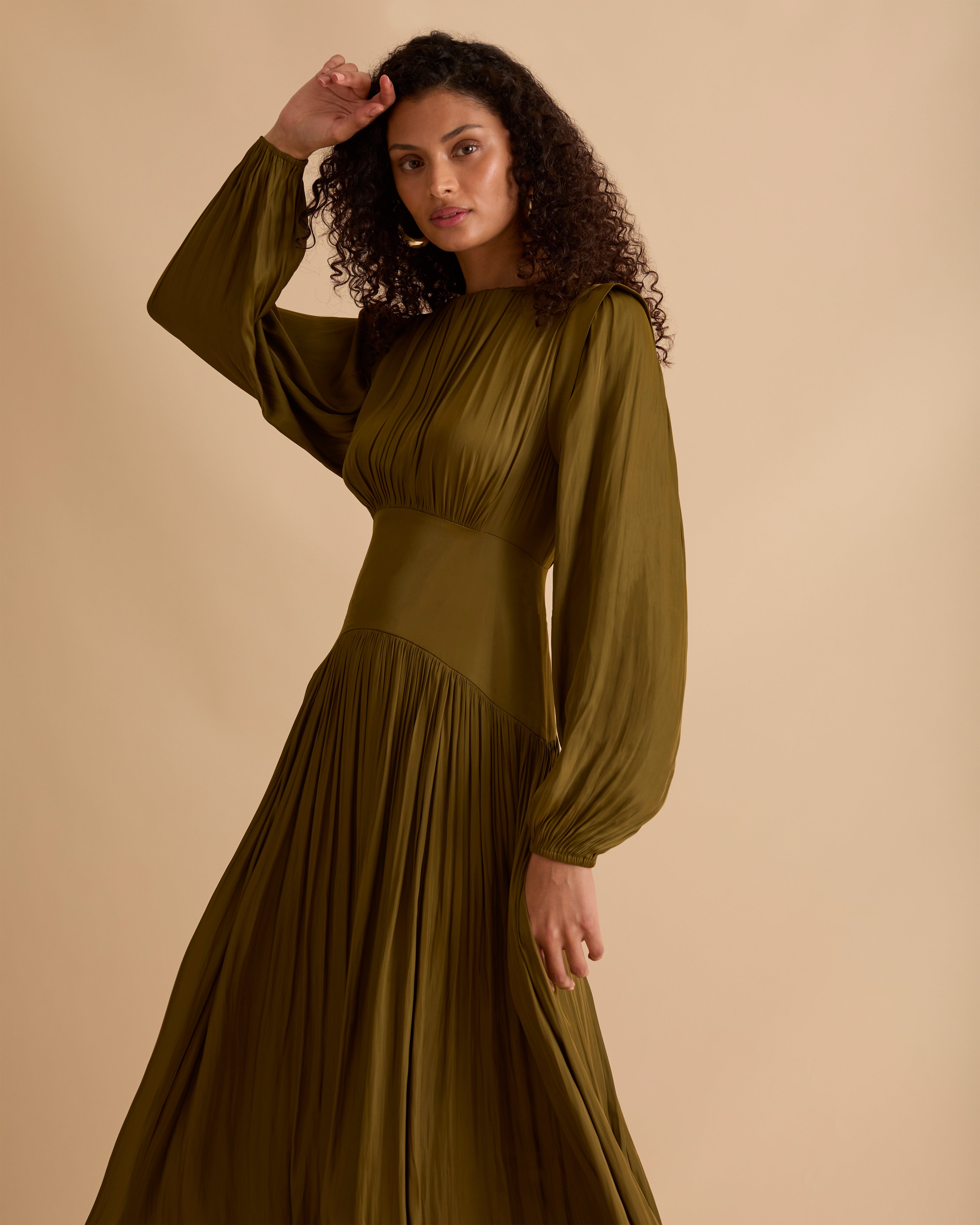 Model wearing Florere olive green maxi dress in billowing sating fabric, with high neck and long sleeves, flattering waist panel and gold zip at the back. Wearing black knee high boots and gold drop earrings against a beige, neutral backdrop