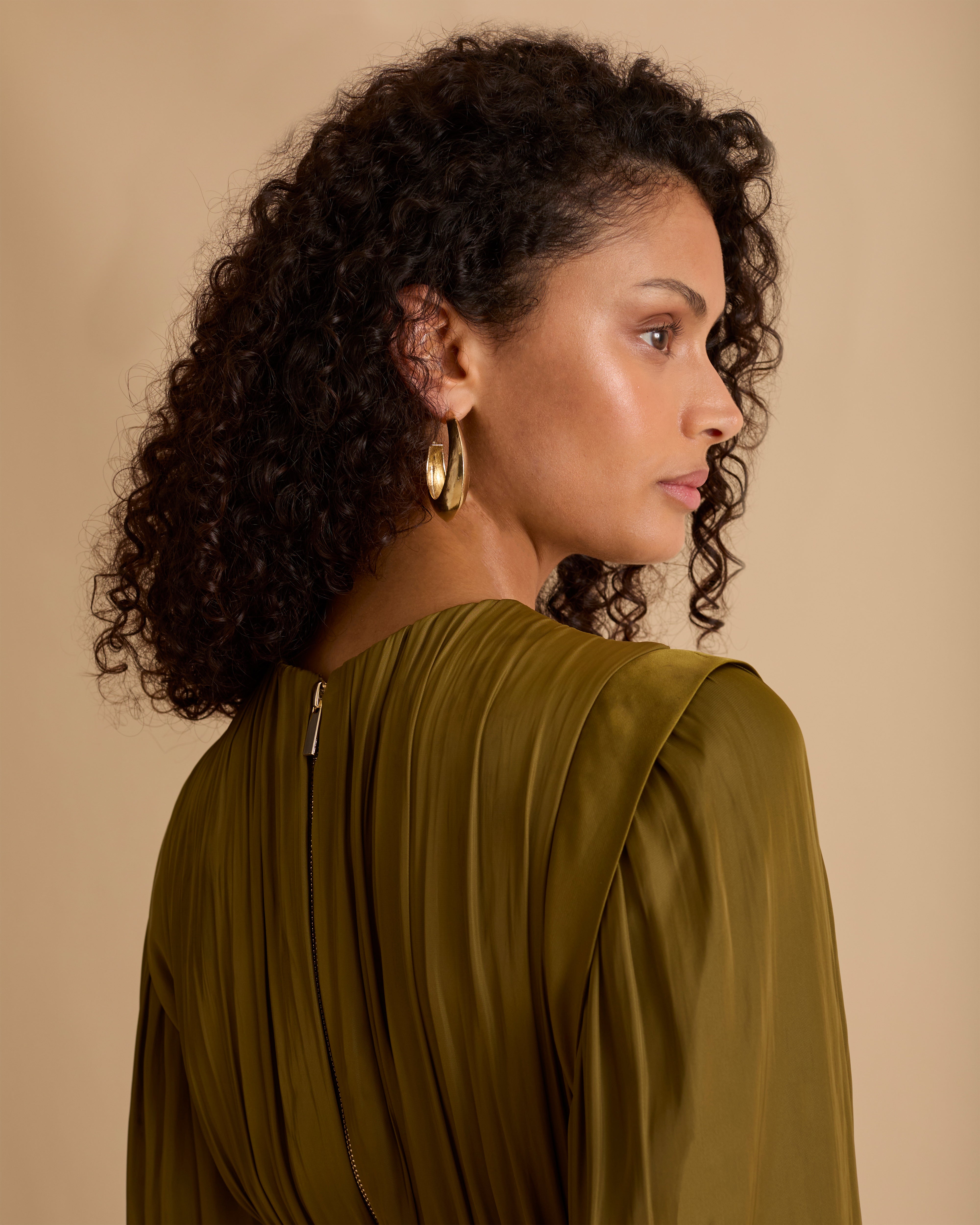 Model wearing Florere olive green maxi dress in billowing sating fabric, with high neck and long sleeves, flattering waist panel and gold zip at the back. Wearing black knee high boots and gold drop earrings against a beige, neutral backdrop