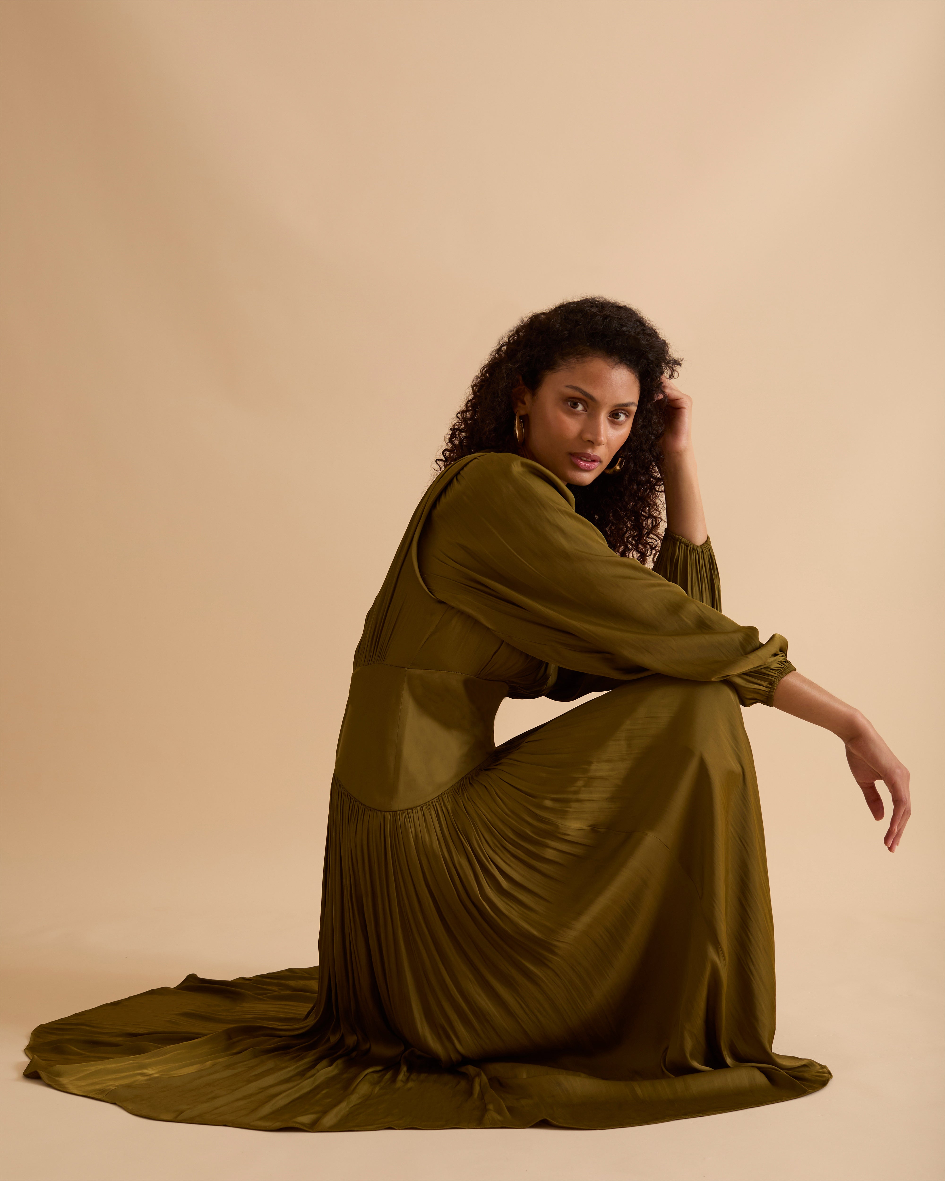 Model wearing Florere olive green maxi dress in billowing sating fabric, with high neck and long sleeves, flattering waist panel and gold zip at the back. Wearing black knee high boots and gold drop earrings against a beige, neutral backdrop