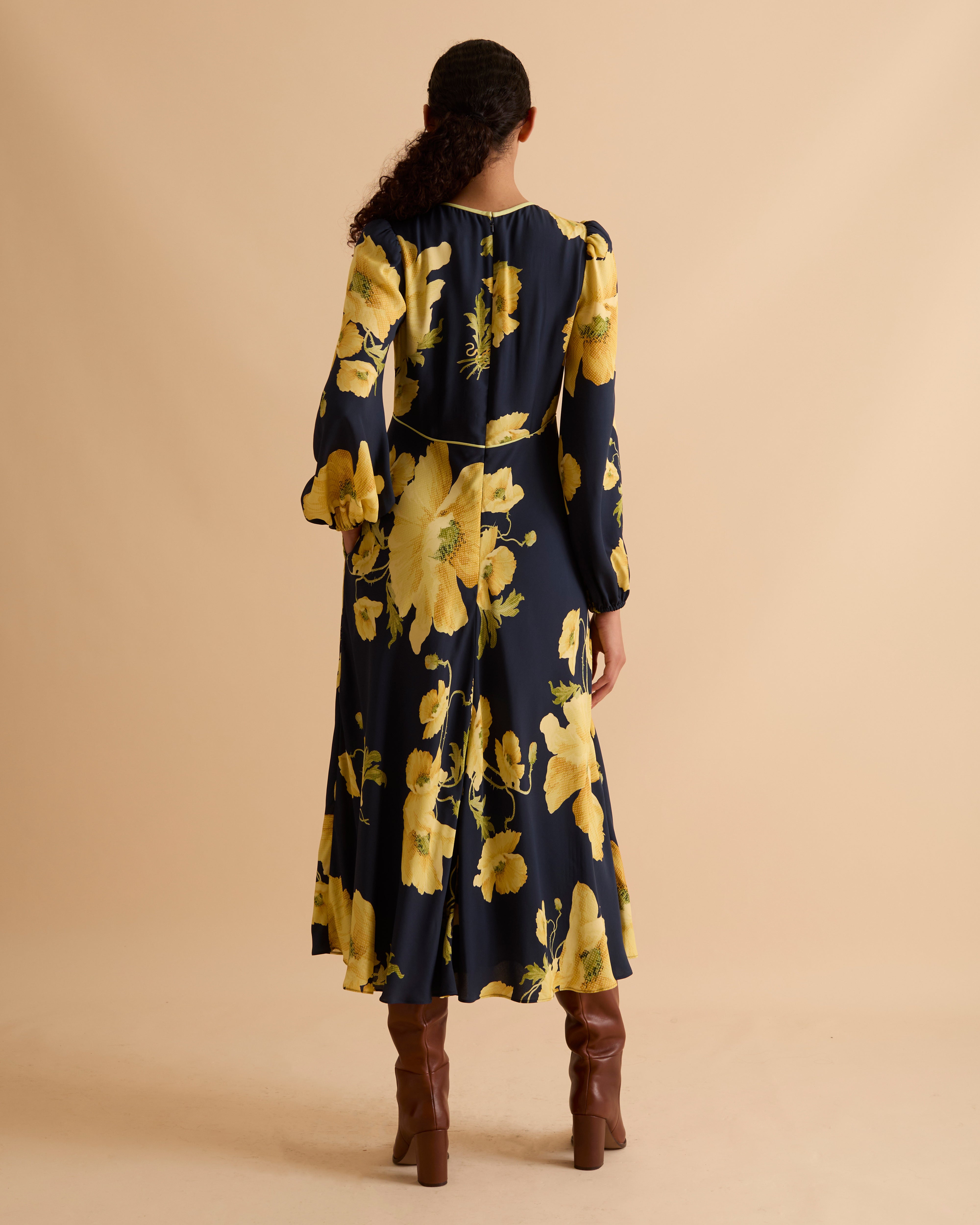 Model wearing Florere floral print occasionwear midi dress. Navy with yellow flowers, high neck dress, with long sleeves, fitted bodice, full skirt with pockets. Wearing tan knee high boots against a beige neutral backdrop