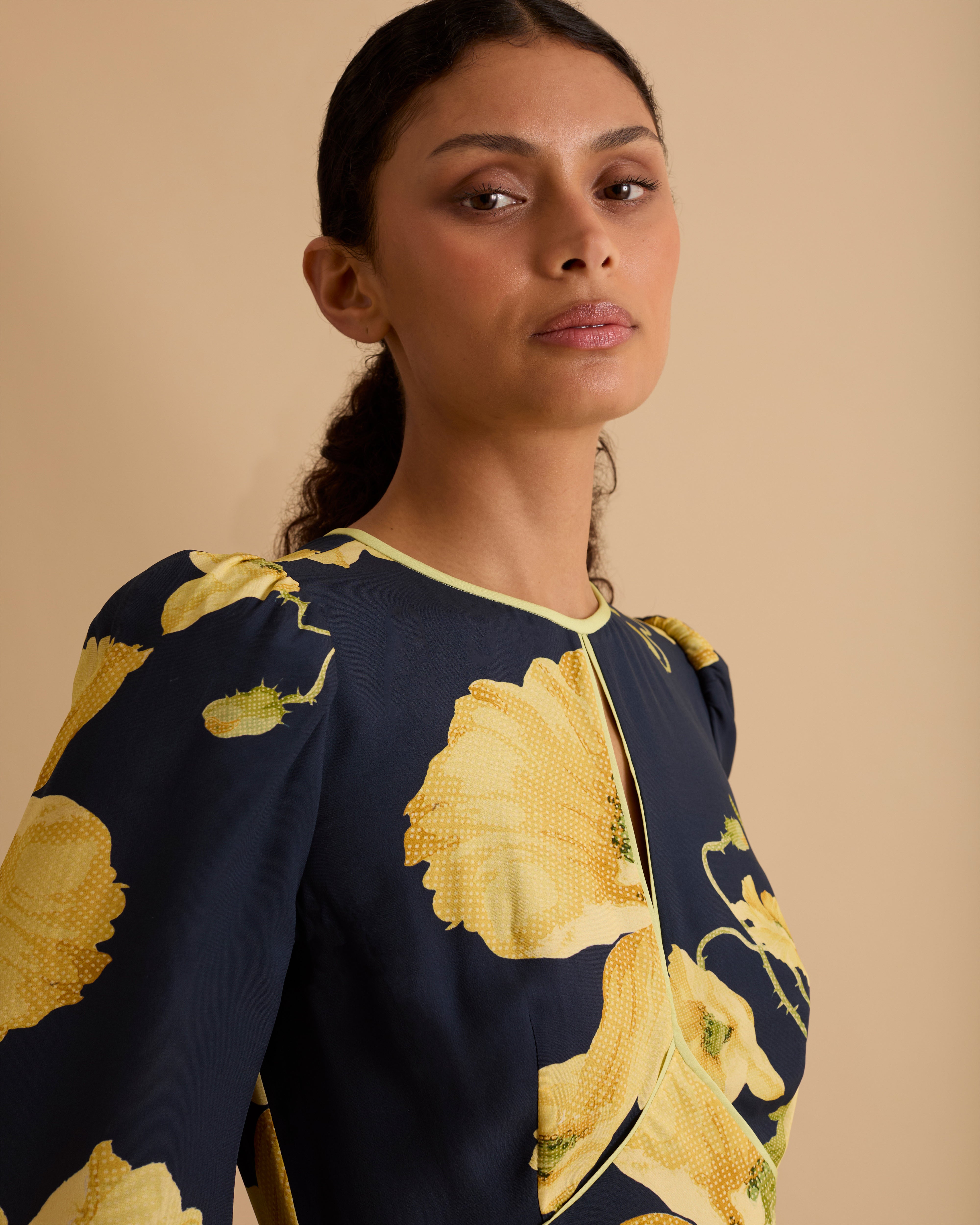 Model wearing Florere floral print occasionwear midi dress. Navy with yellow flowers, high neck dress, with long sleeves, fitted bodice, full skirt with pockets. Wearing tan knee high boots against a beige neutral backdrop