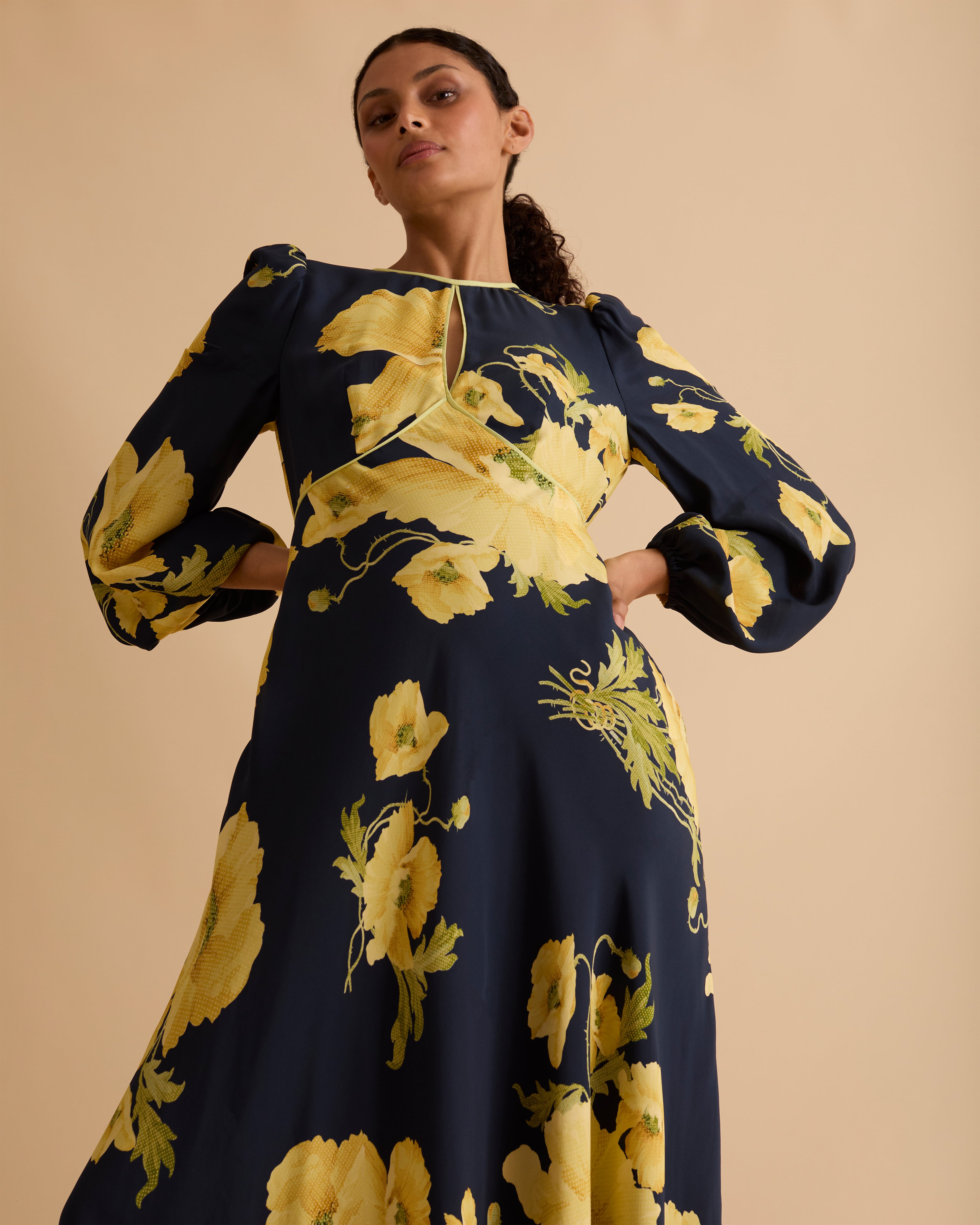 Model wearing Florere floral print occasionwear midi dress. Navy with yellow flowers, high neck dress, with long sleeves, fitted bodice, full skirt with pockets. Wearing tan knee high boots against a beige neutral backdrop