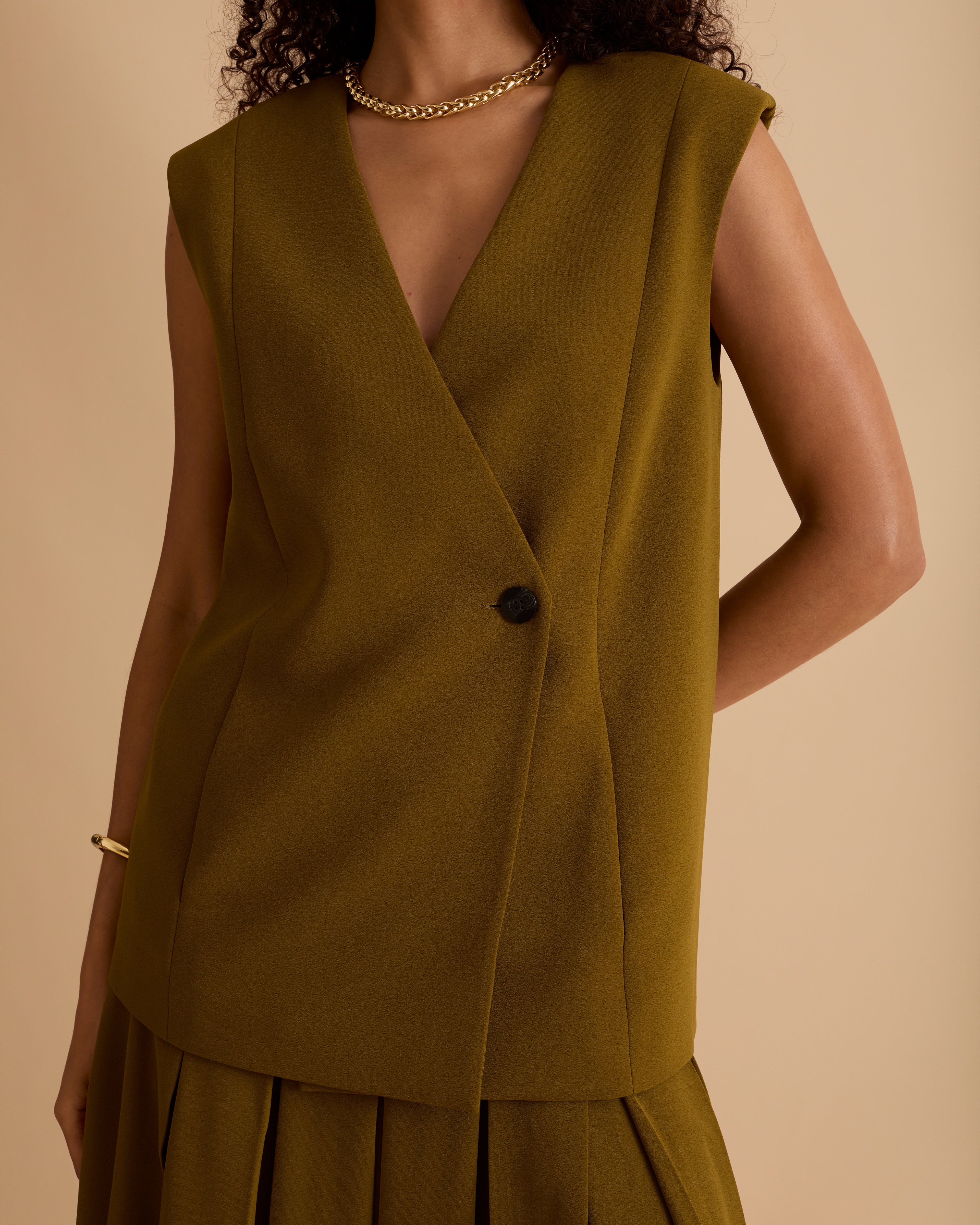 Model wearing Florere sleeveless loose fit waistcoat in Olive green, wearing black boots and matching pleated midi skirt with gold chunky necklace on a beige neutral backdrop