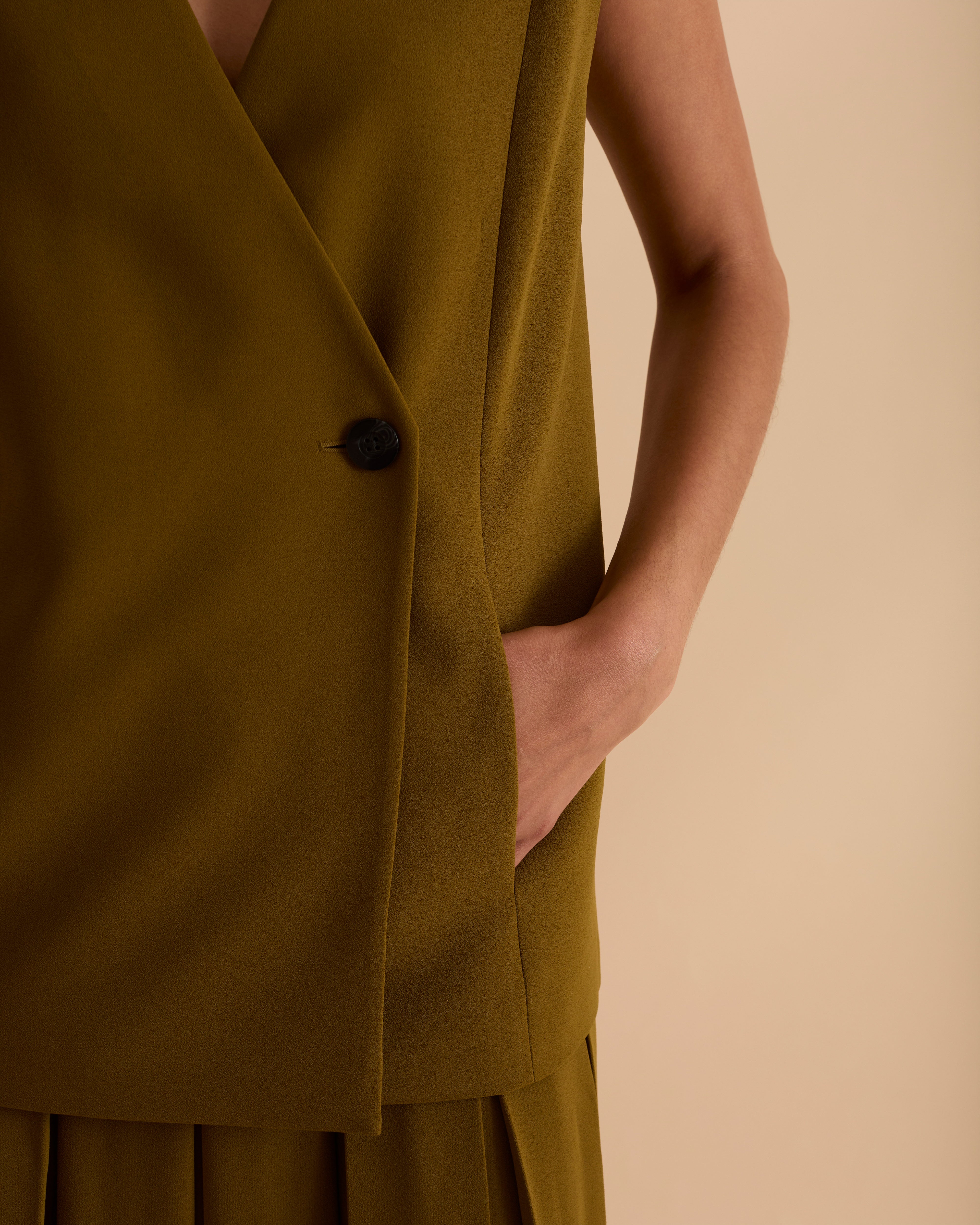Model wearing Florere sleeveless loose fit waistcoat in Olive green, wearing black boots and matching pleated midi skirt with gold chunky necklace on a beige neutral backdrop