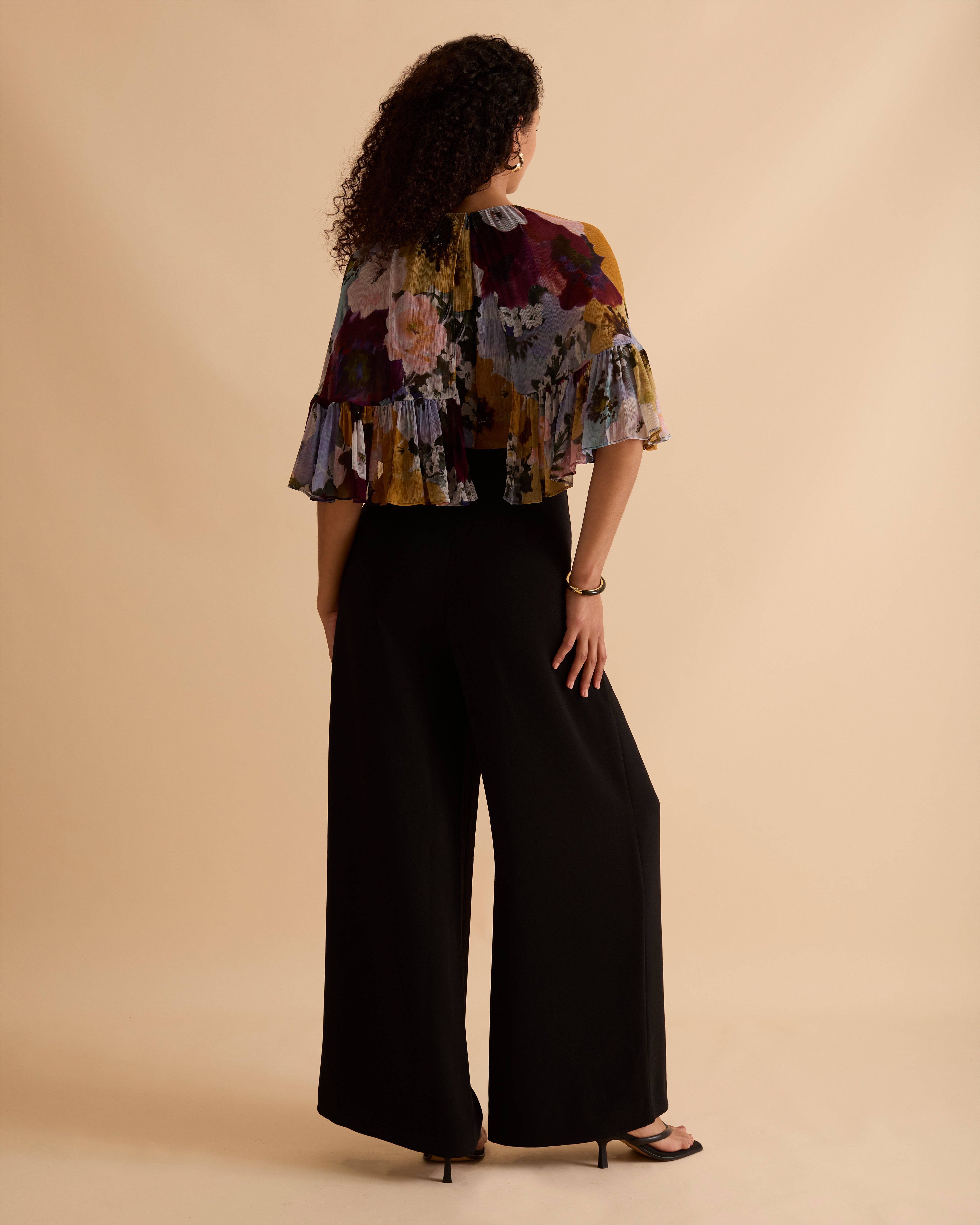 Model wearing Florere jumpsuit, with tailored black trouser and lightweight floral printed cape top, with high neck and covered arms. Against a beige neutral backdrop