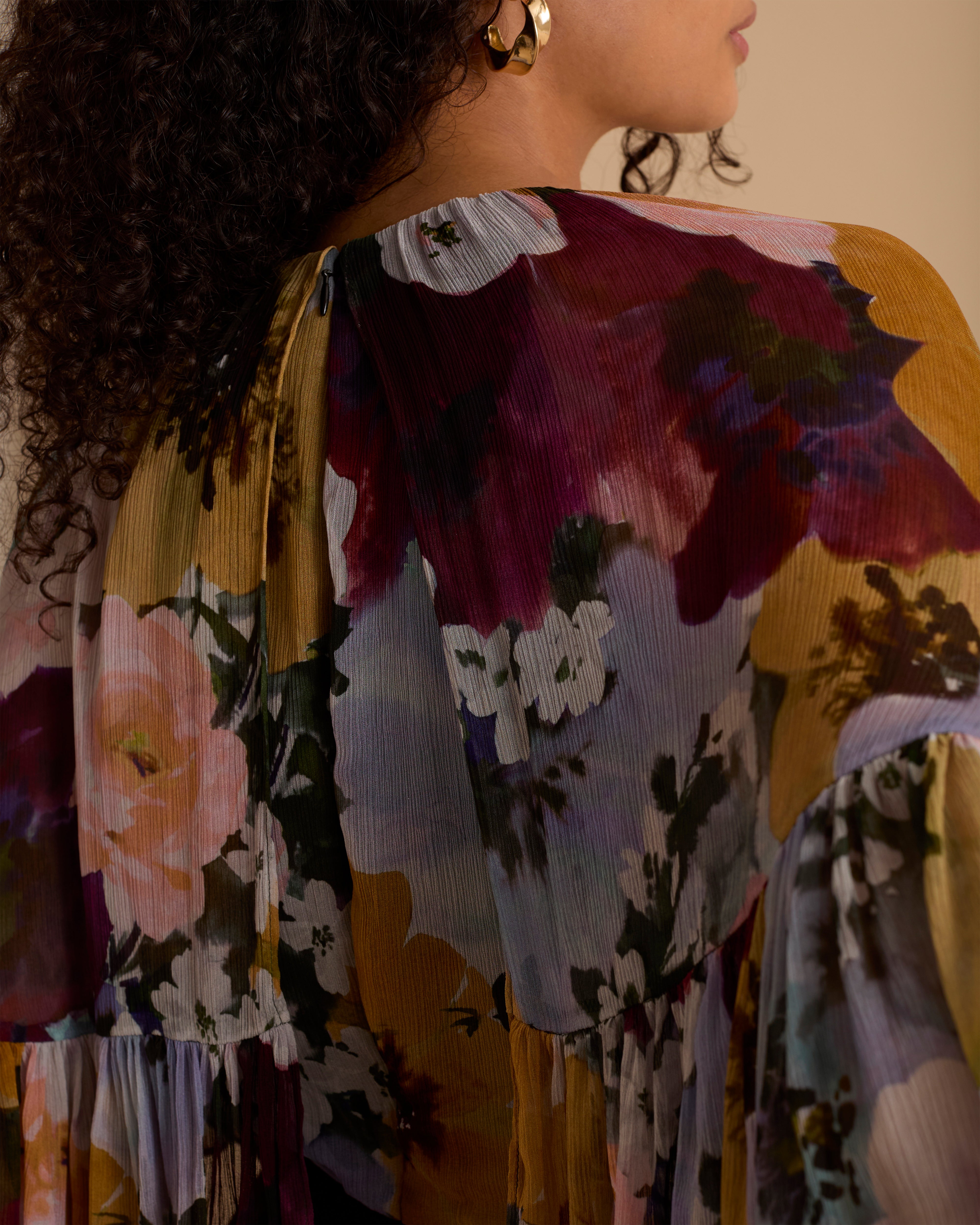 Model wearing Florere jumpsuit, with tailored black trouser and lightweight floral printed cape top, with high neck and covered arms. Against a beige neutral backdrop
