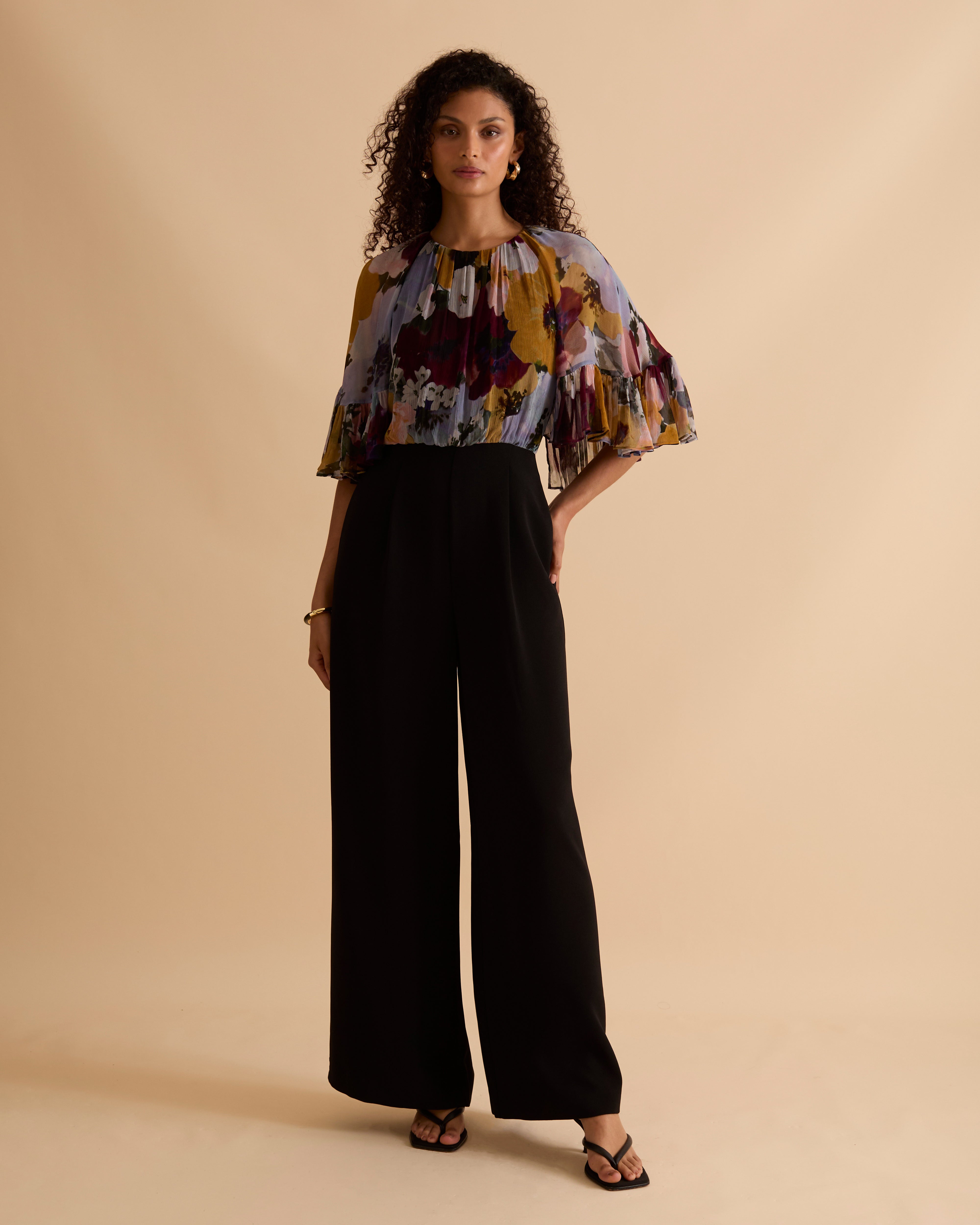 Model wearing Florere jumpsuit, with tailored black trouser and lightweight floral printed cape top, with high neck and covered arms. Against a beige neutral backdrop