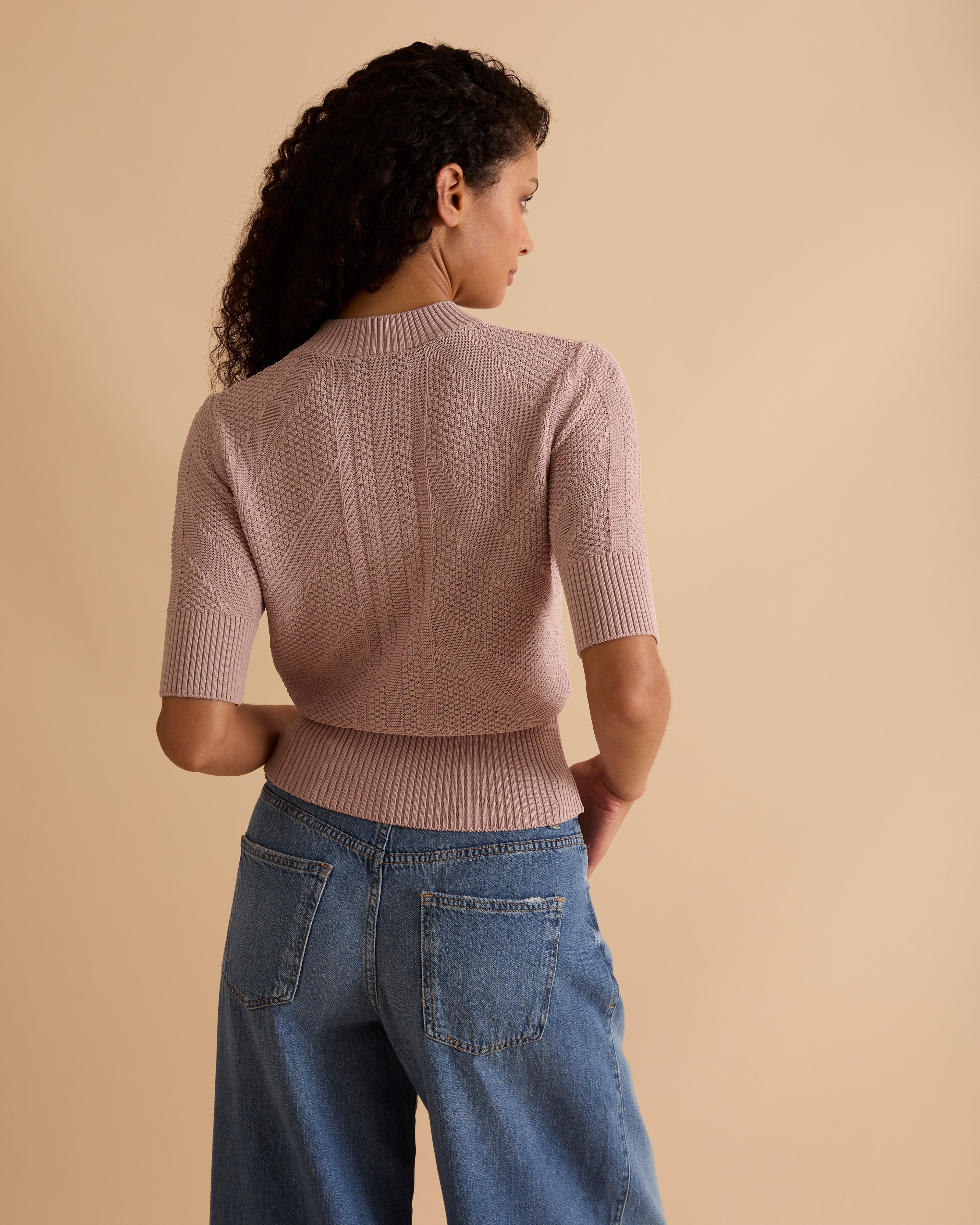 Model wearing Florere light pink, pointelle knit short sleeve lighweight jumper, with crew neck, worn with light blue wide legged jeans, against a beige neutral backdrop