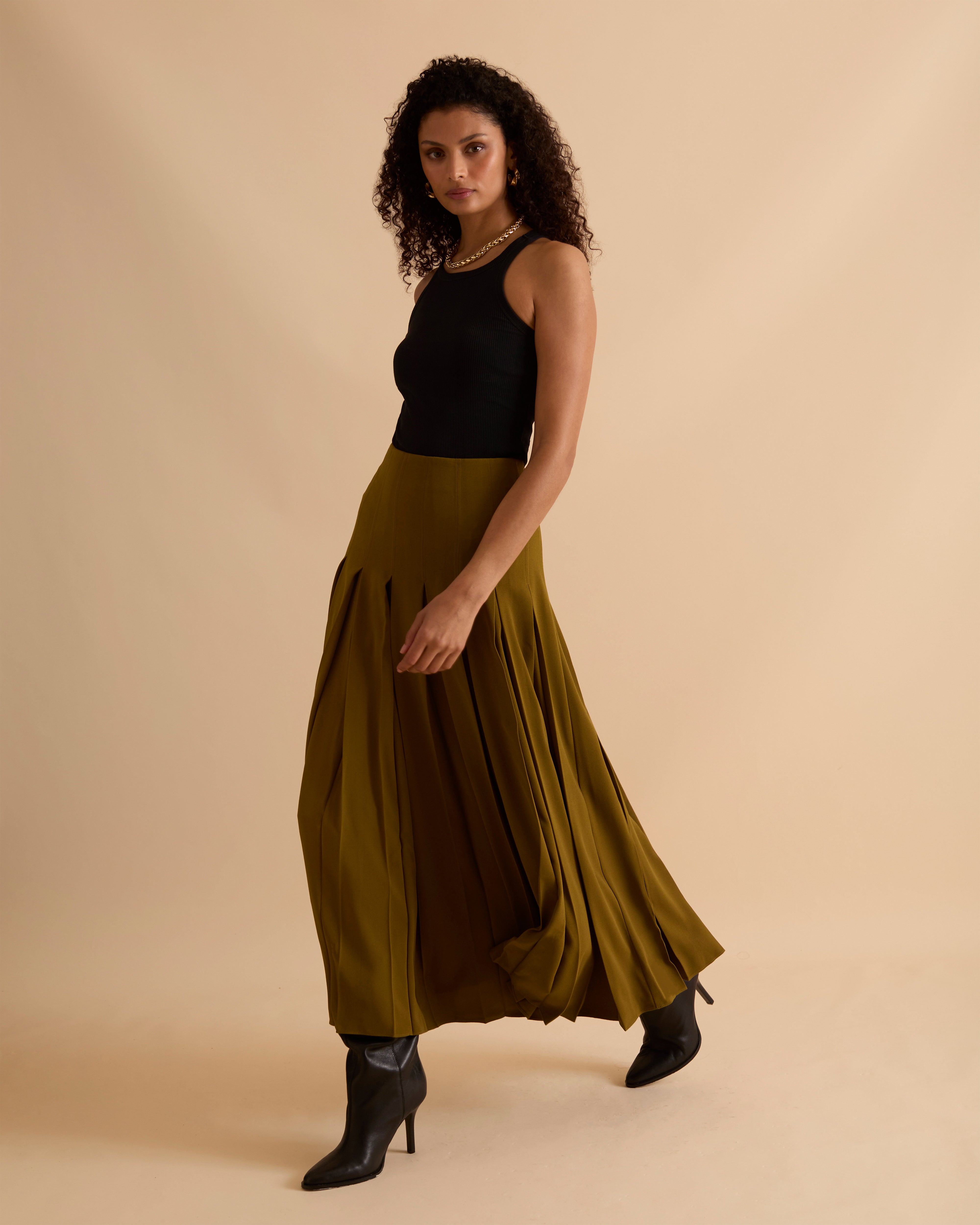 Model wearing Florere deep pleated midi skirt in olive green, wearing black boots and matching sleeveless waistcoat with gold chunky necklace on a beige neutral backdrop