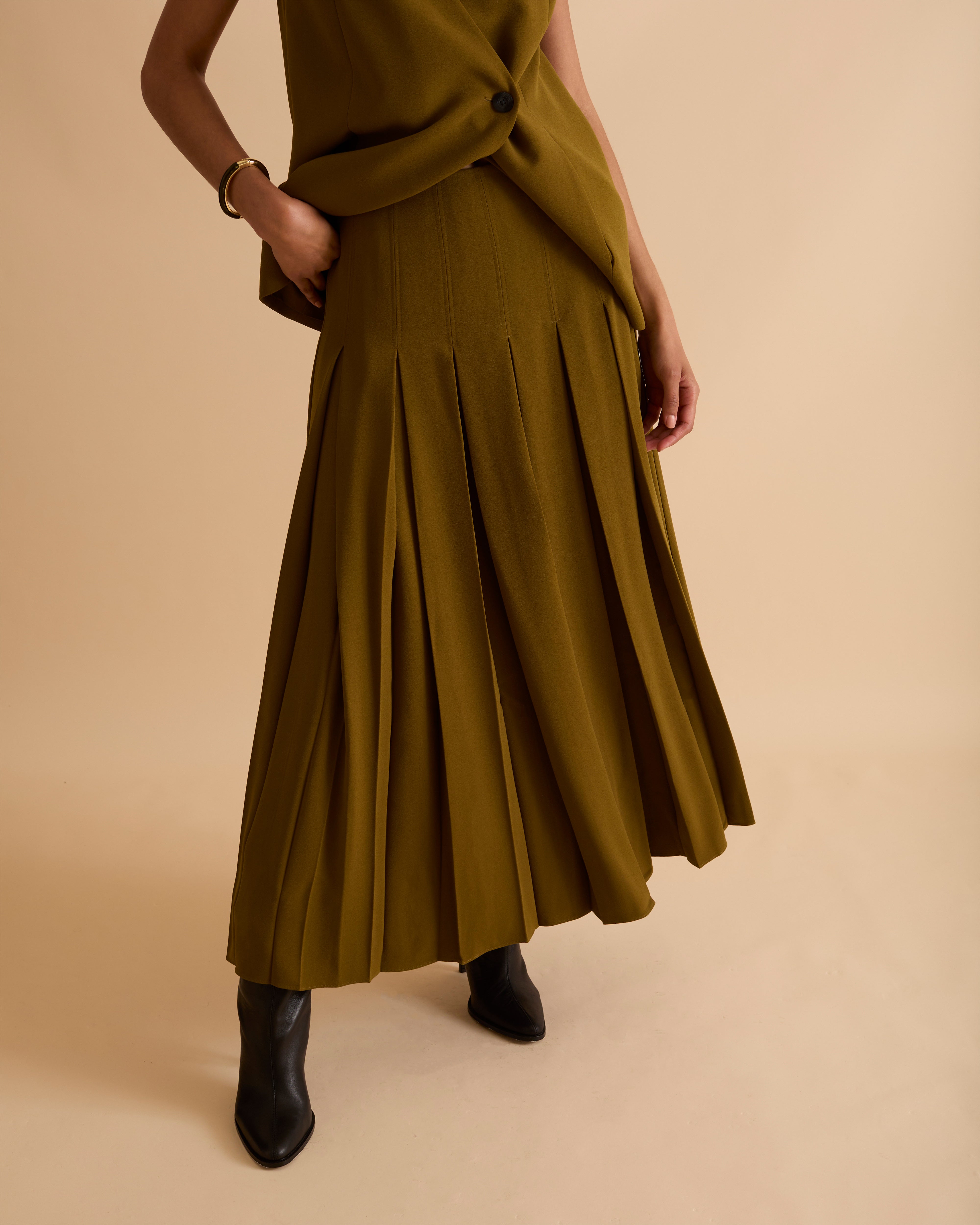 Model wearing Florere deep pleated midi skirt in olive green, wearing black boots and matching sleeveless waistcoat with gold chunky necklace on a beige neutral backdrop